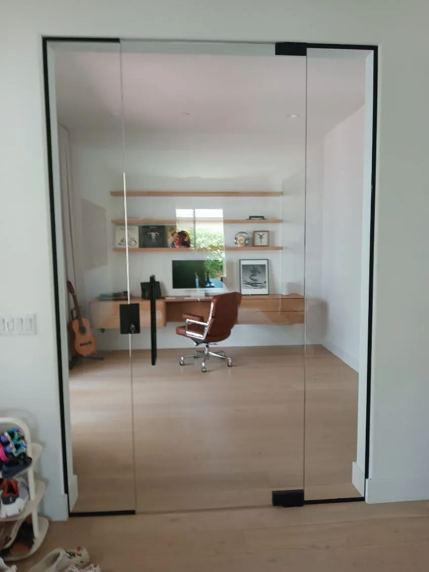 Modern home office seen through mirrored closet doors, with desk, shelves, chair, and guitar.