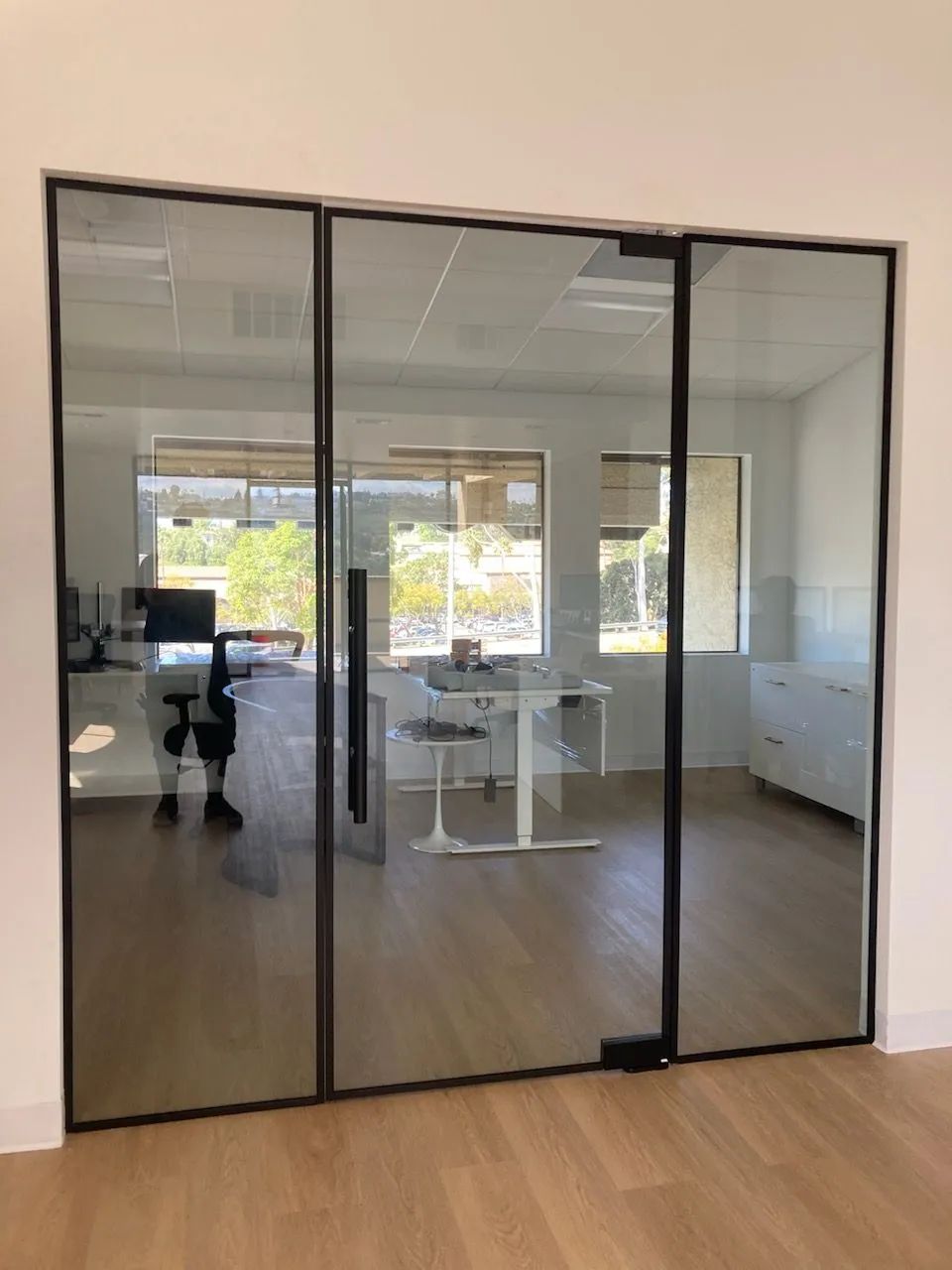 Glass-walled office meeting room with light wood floors and white desks, viewed through a doorway