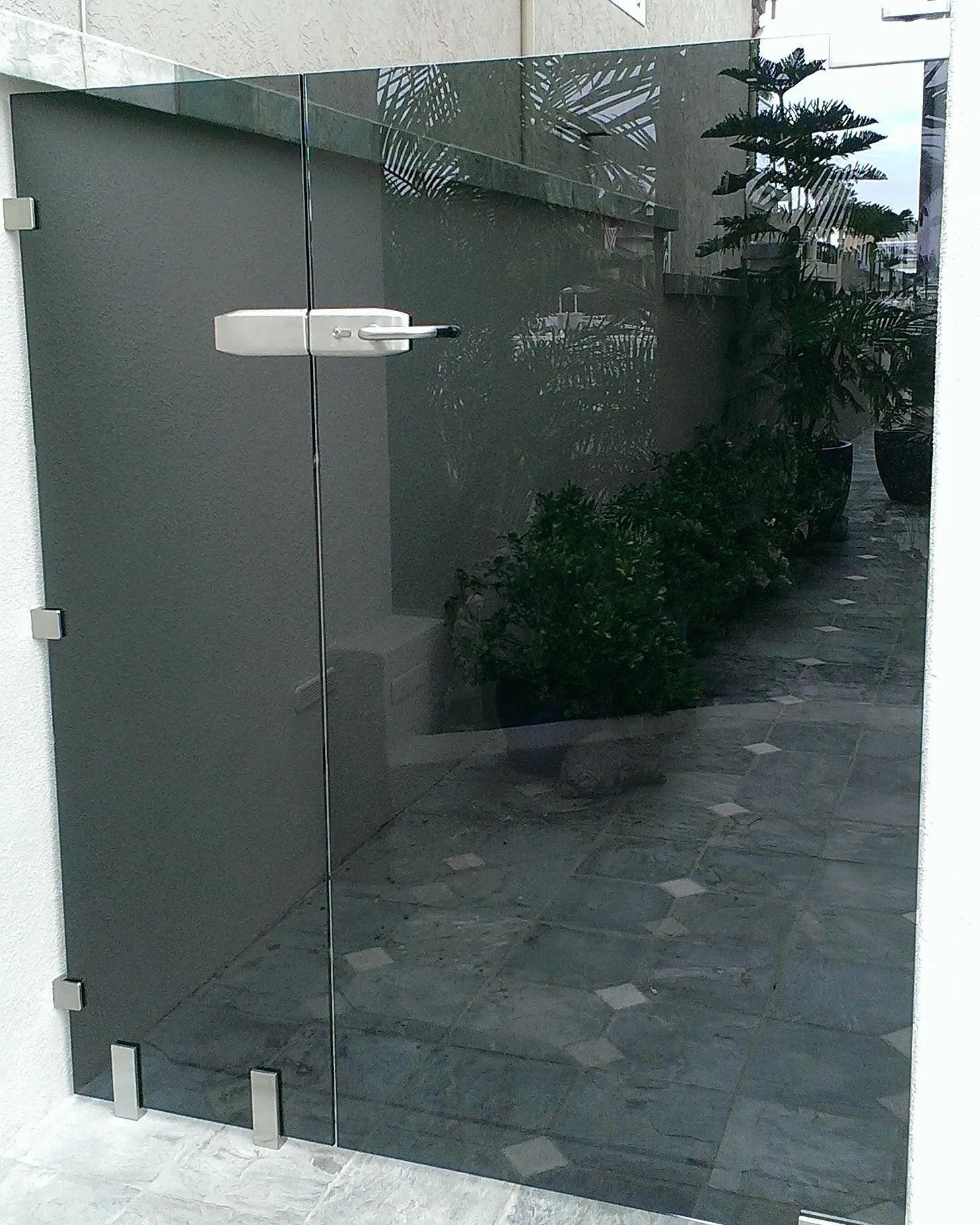 Glass entrance door with frosted panel and metal handle beside a walkway with greenery