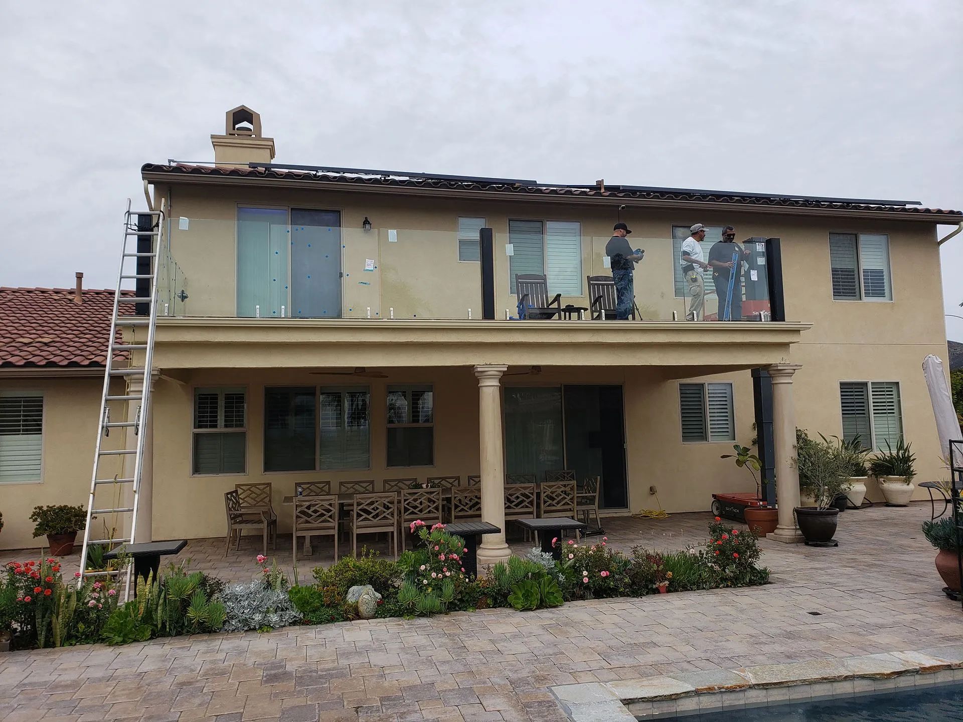 Two-story beige house with a covered porch, ladder on the left, and workers on the upper balcony.