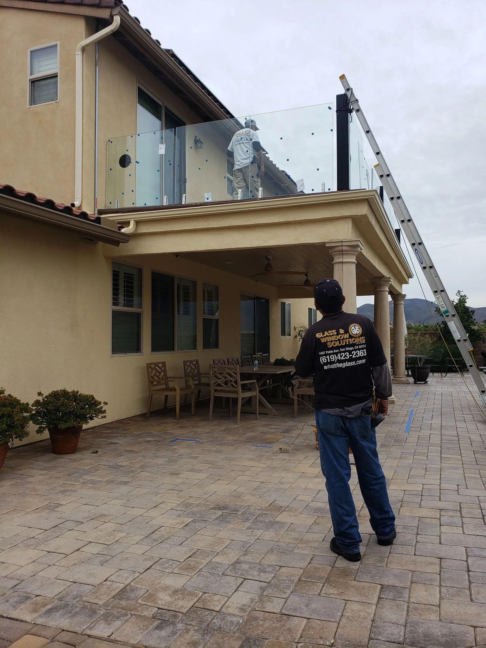 Person standing in a paved courtyard beside a beige house with glass balcony railings and an outdoor ladder