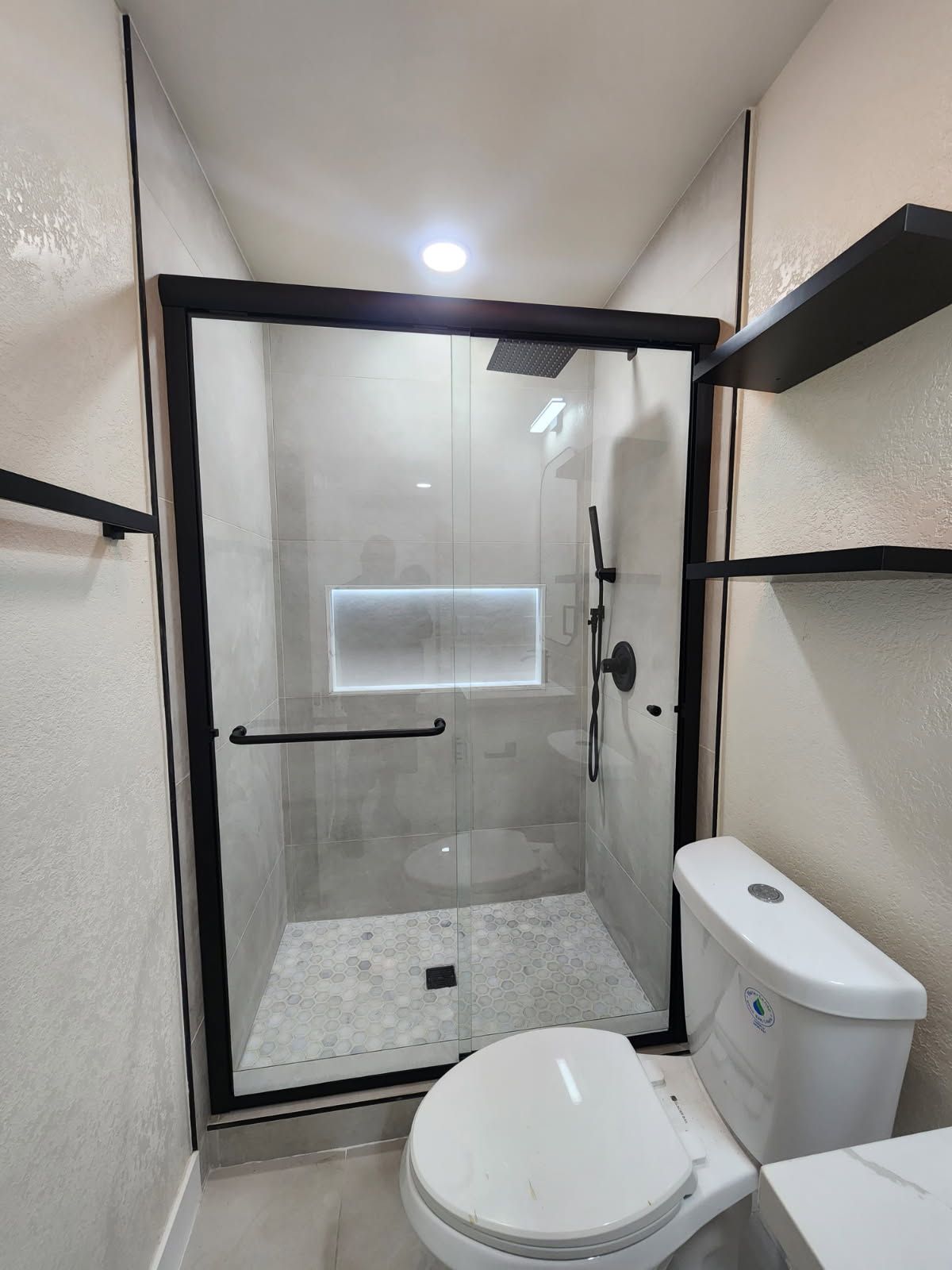 Modern bathroom with glass shower, toilet, and black shelving on beige walls