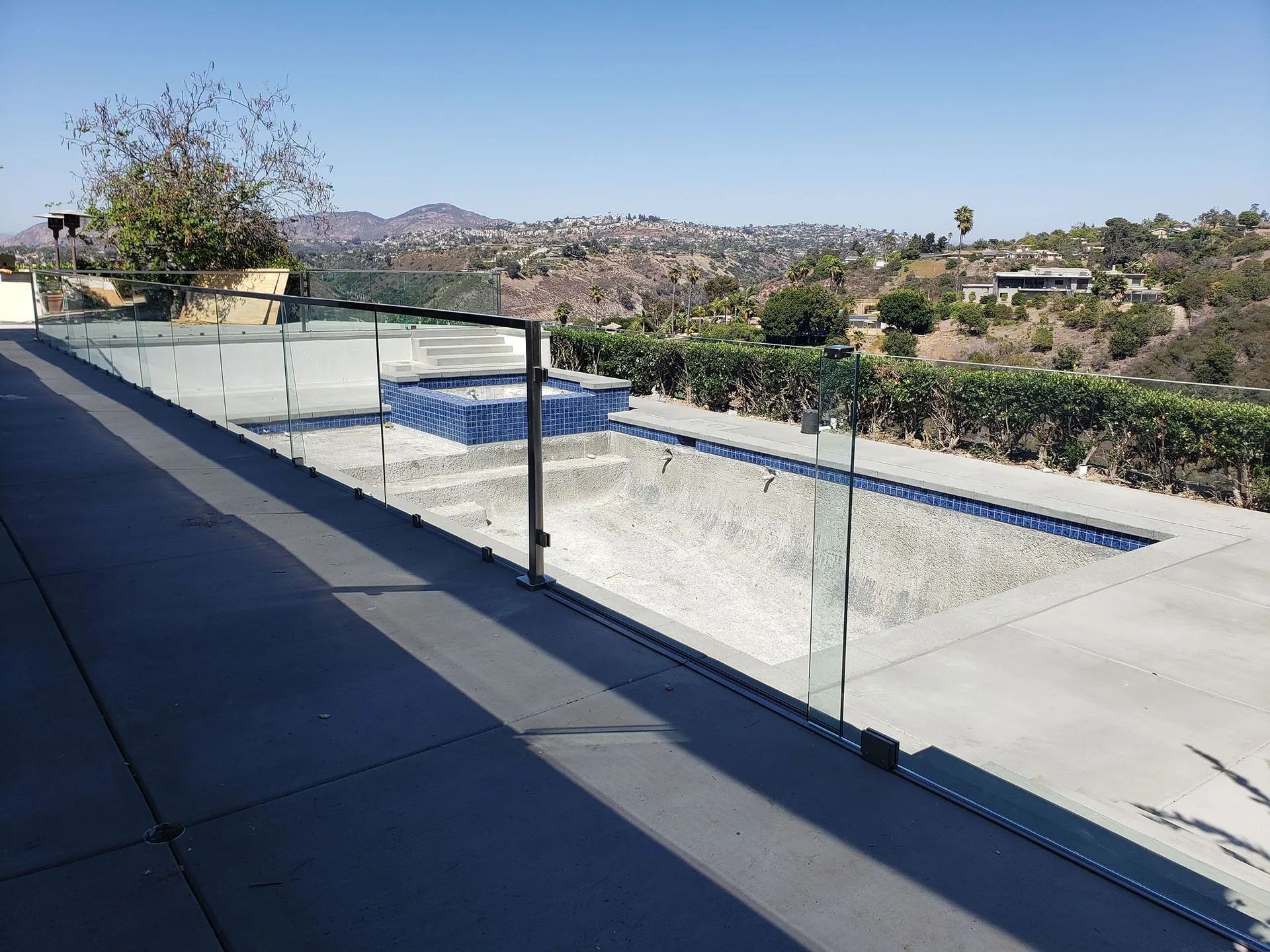 Backyard pool with glass railing, concrete deck, and hillside views under a clear blue sky