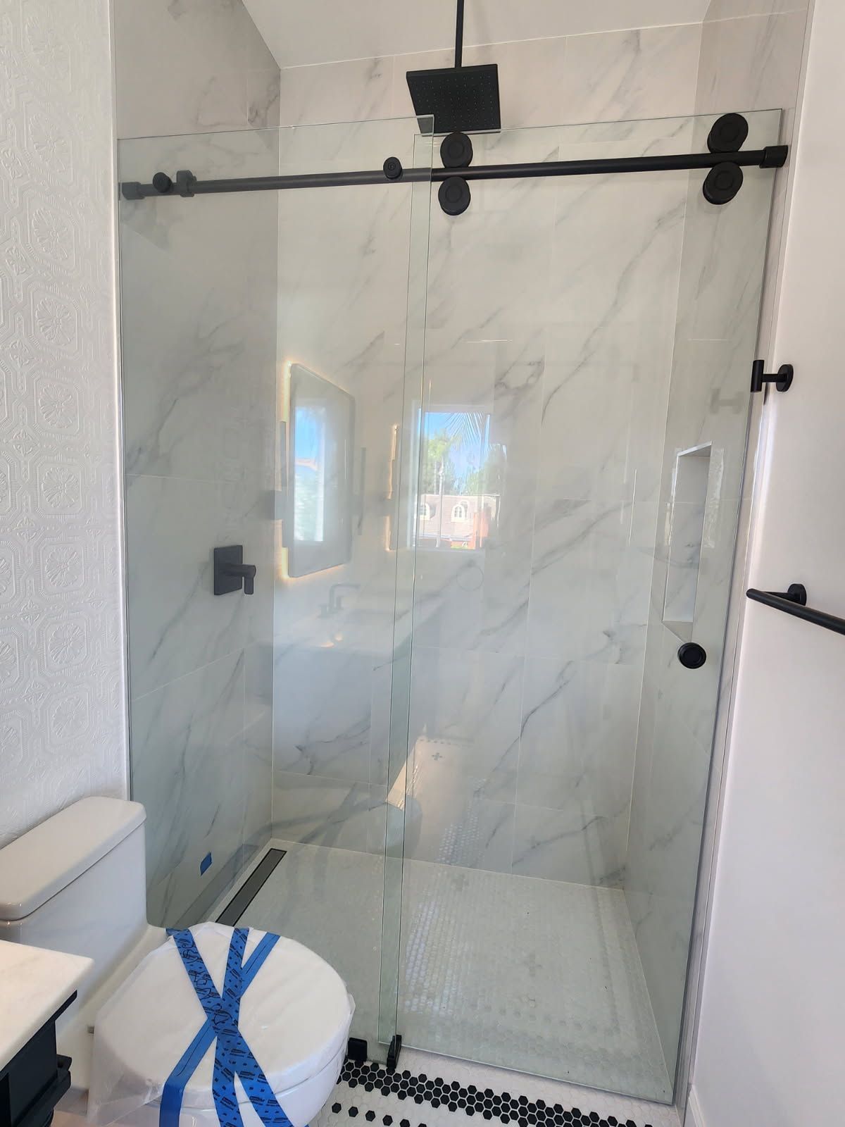 Modern bathroom with glass shower enclosure, marble-look tile, and a toilet marked with blue tape.