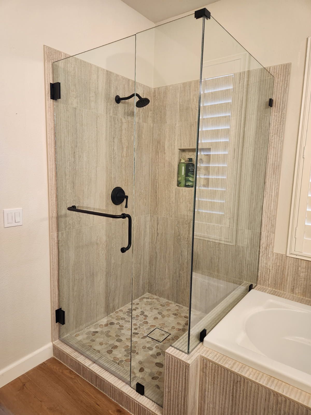 Glass walk-in shower with black fixtures and beige tile beside a white bathtub