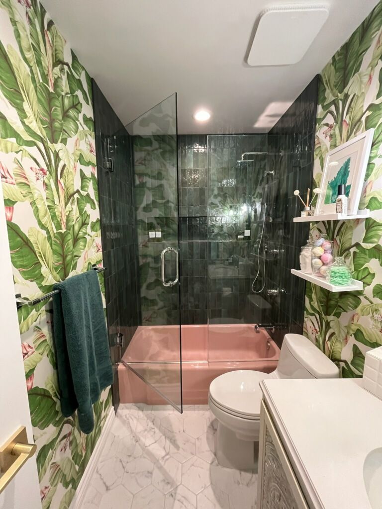 Narrow bathroom with leafy wallpaper, glass shower, pink tile, marble floor, and white vanity.