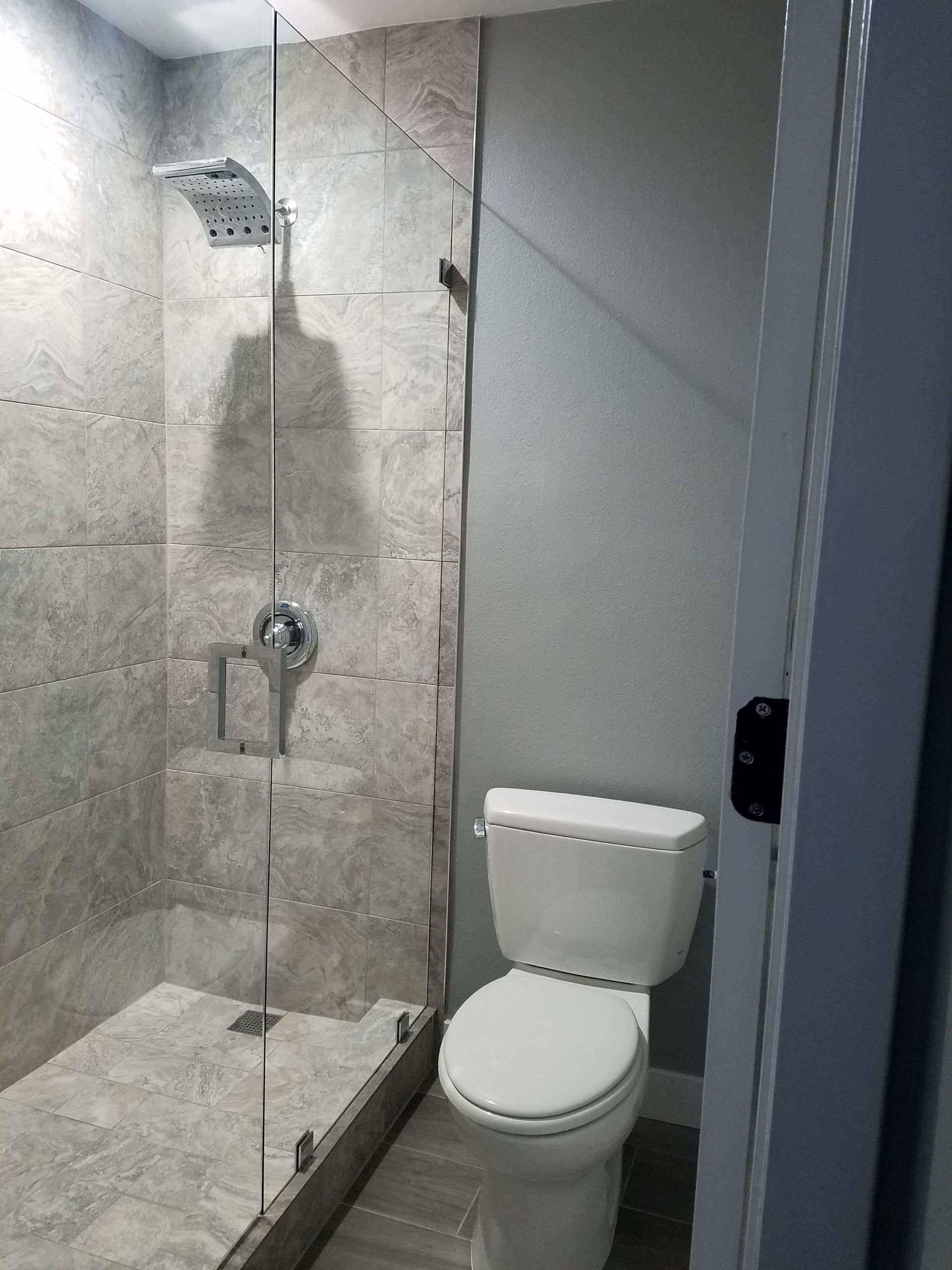 Small gray-tiled bathroom with a toilet and walk-in shower behind a glass partition.