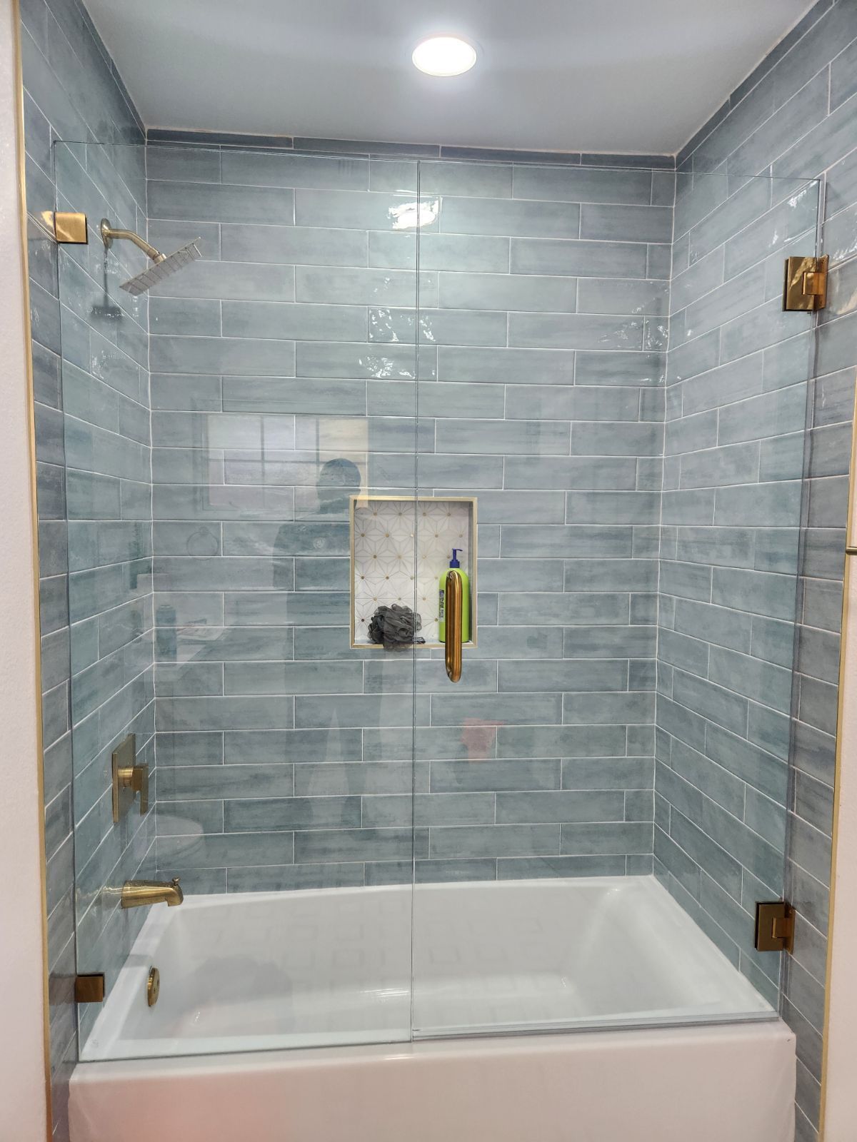 Walk-in shower with gray tile walls, white tub base, and brass fixtures under a ceiling light