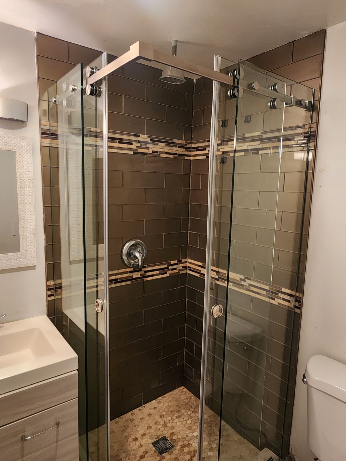 Modern glass walk-in shower with chrome fixtures, dark tile walls, and pebble stone floor in a bathroom
