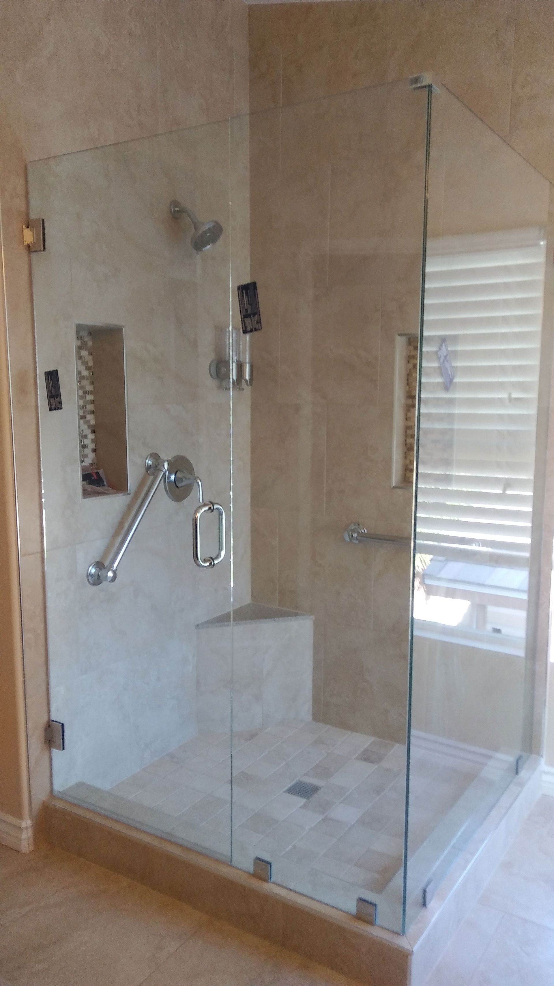 Glass-enclosed walk-in shower with beige tile, chrome fixtures, built-in bench, and window blinds.