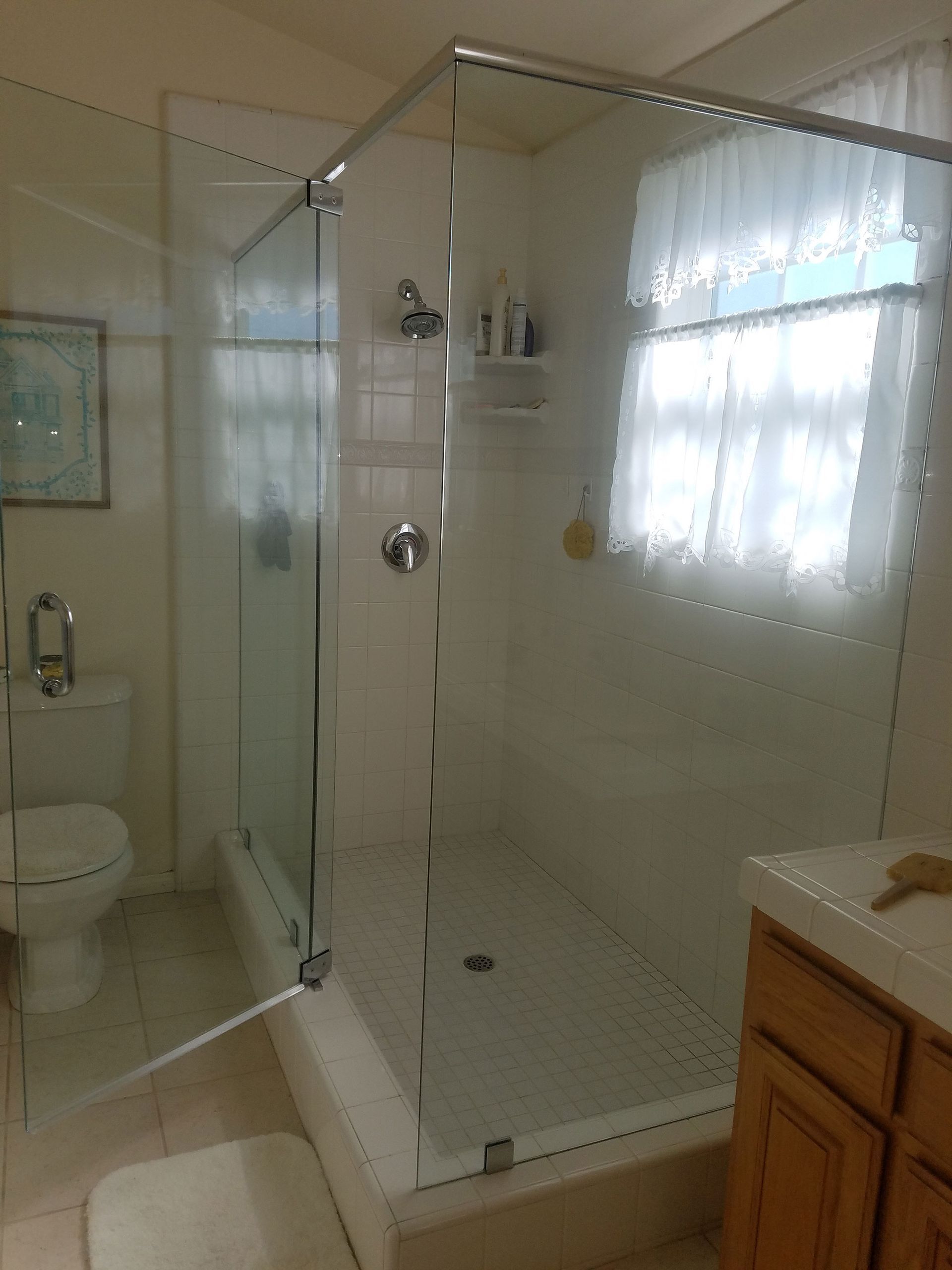 Bright bathroom with glass shower enclosure, toilet, and window over a wooden vanity.