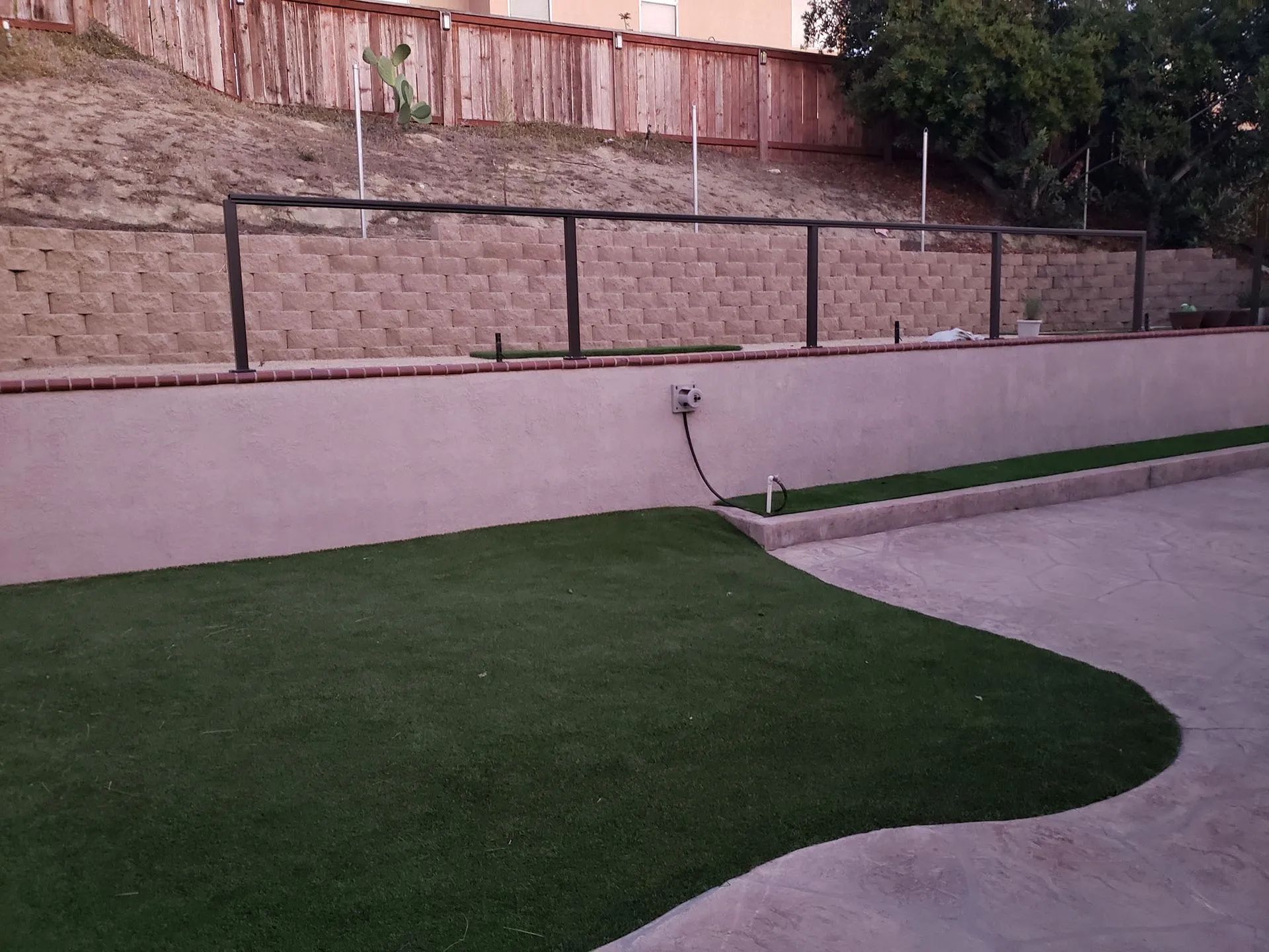 Curved concrete patio beside green lawn, pink retaining wall, and stone fence in a backyard