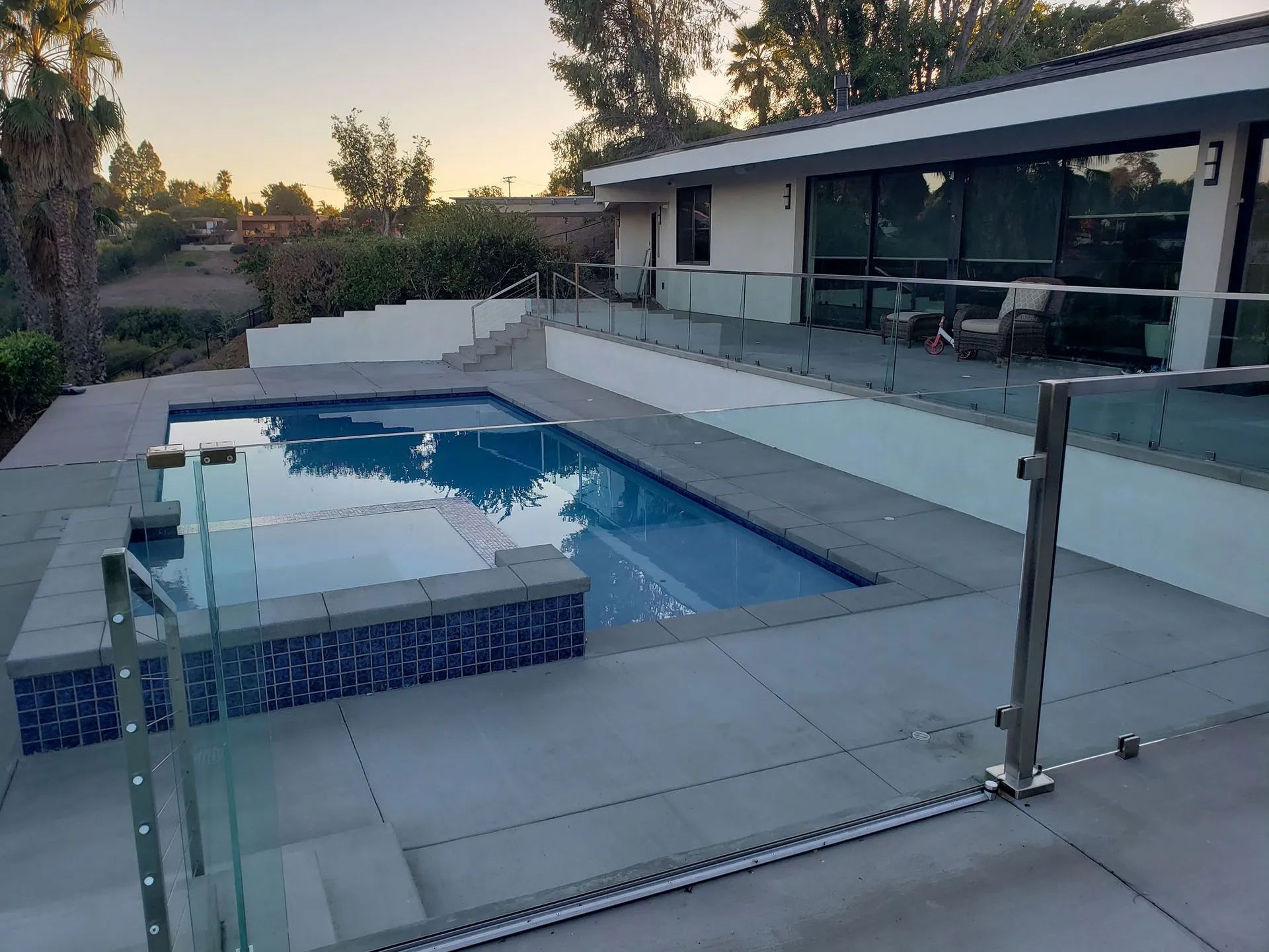 Modern backyard patio with a blue swimming pool, glass fence, and a house at sunset