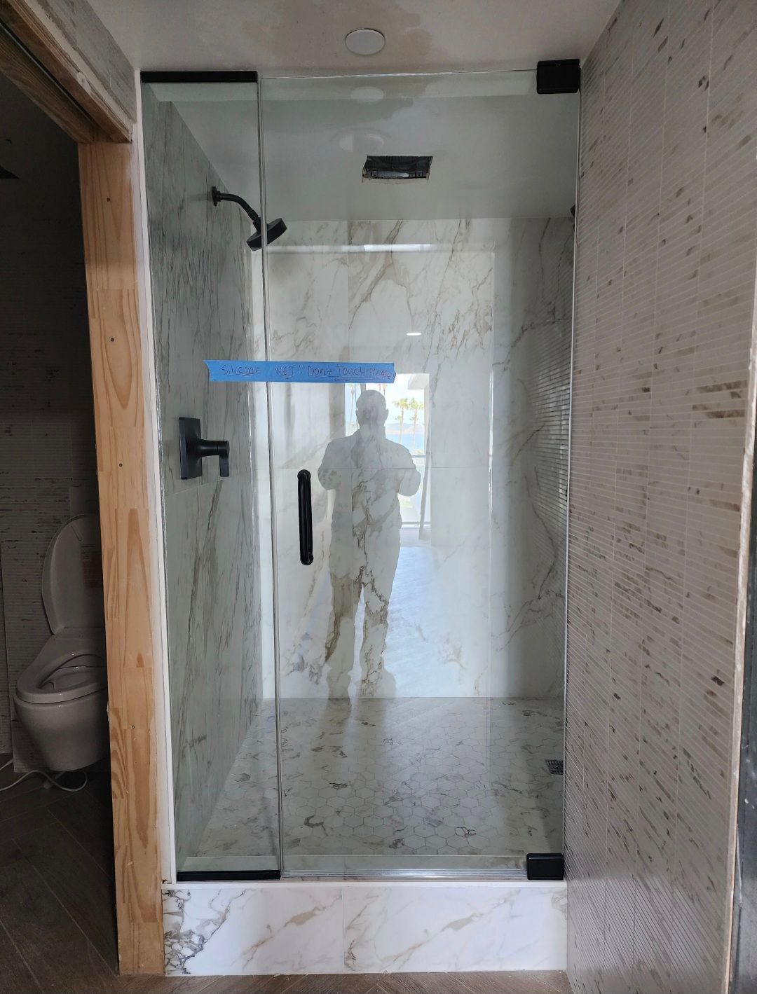 Glass shower enclosure in a marble-tiled bathroom with a black handle and blue tape across the door.