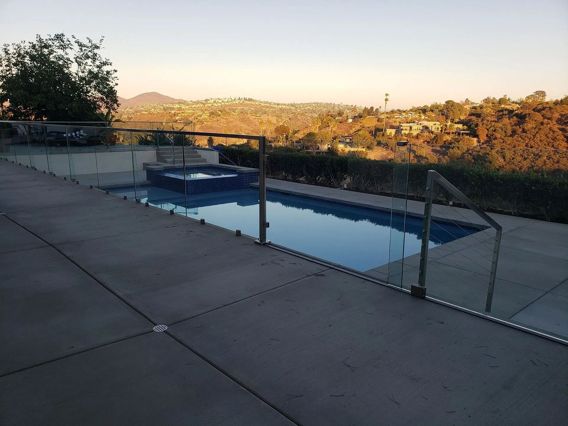 Backyard patio with a rectangular pool, glass fence, and hills in warm sunset light