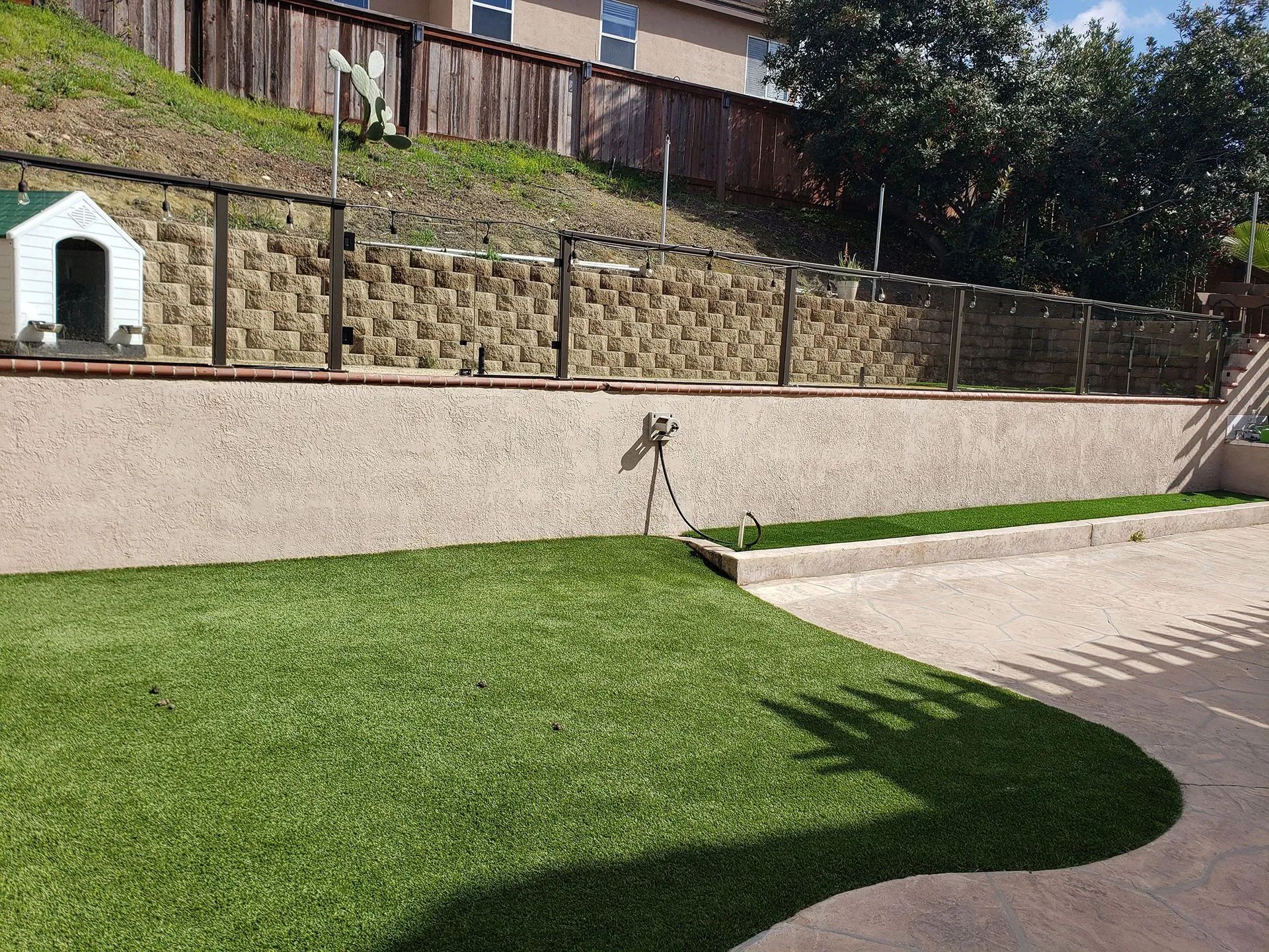 Backyard lawn with stone retaining wall, small pet house, and patio edge in sunlight