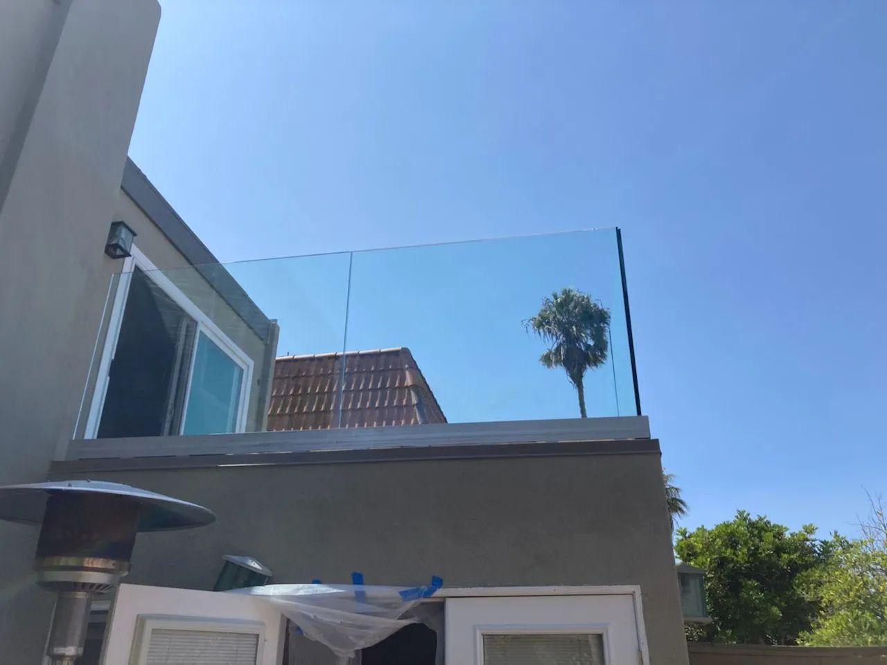 Rooftop patio with glass railing, gray walls, blue sky, and a small palm tree in the distance