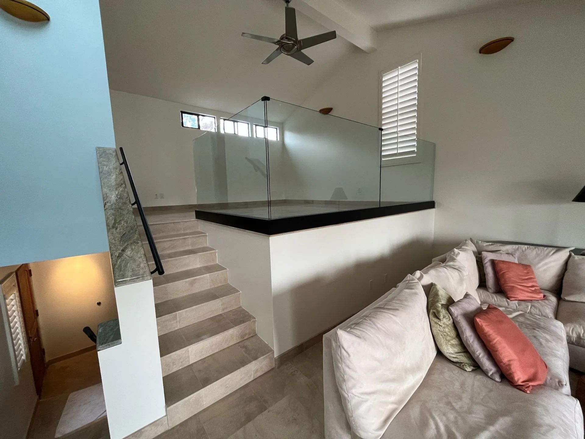 Bright loft living room with stairs, ceiling fan, raised platform, and a gray sofa with coral pillows