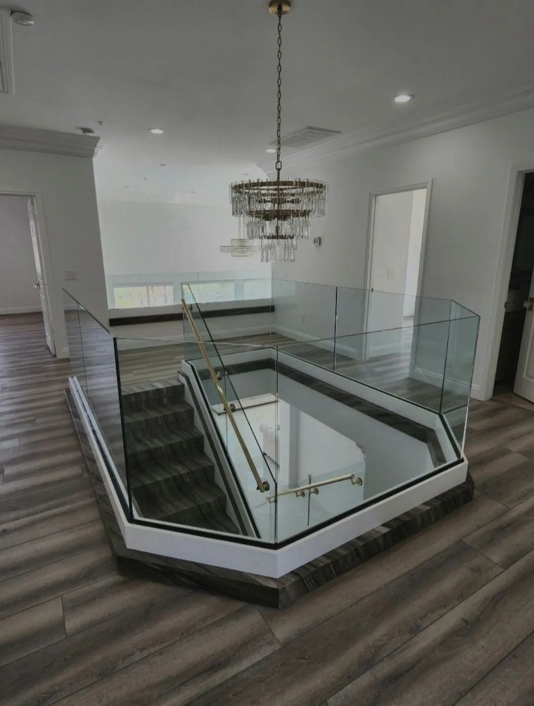 Modern foyer with glass-railed staircase and chandelier, white walls, and wood flooring.