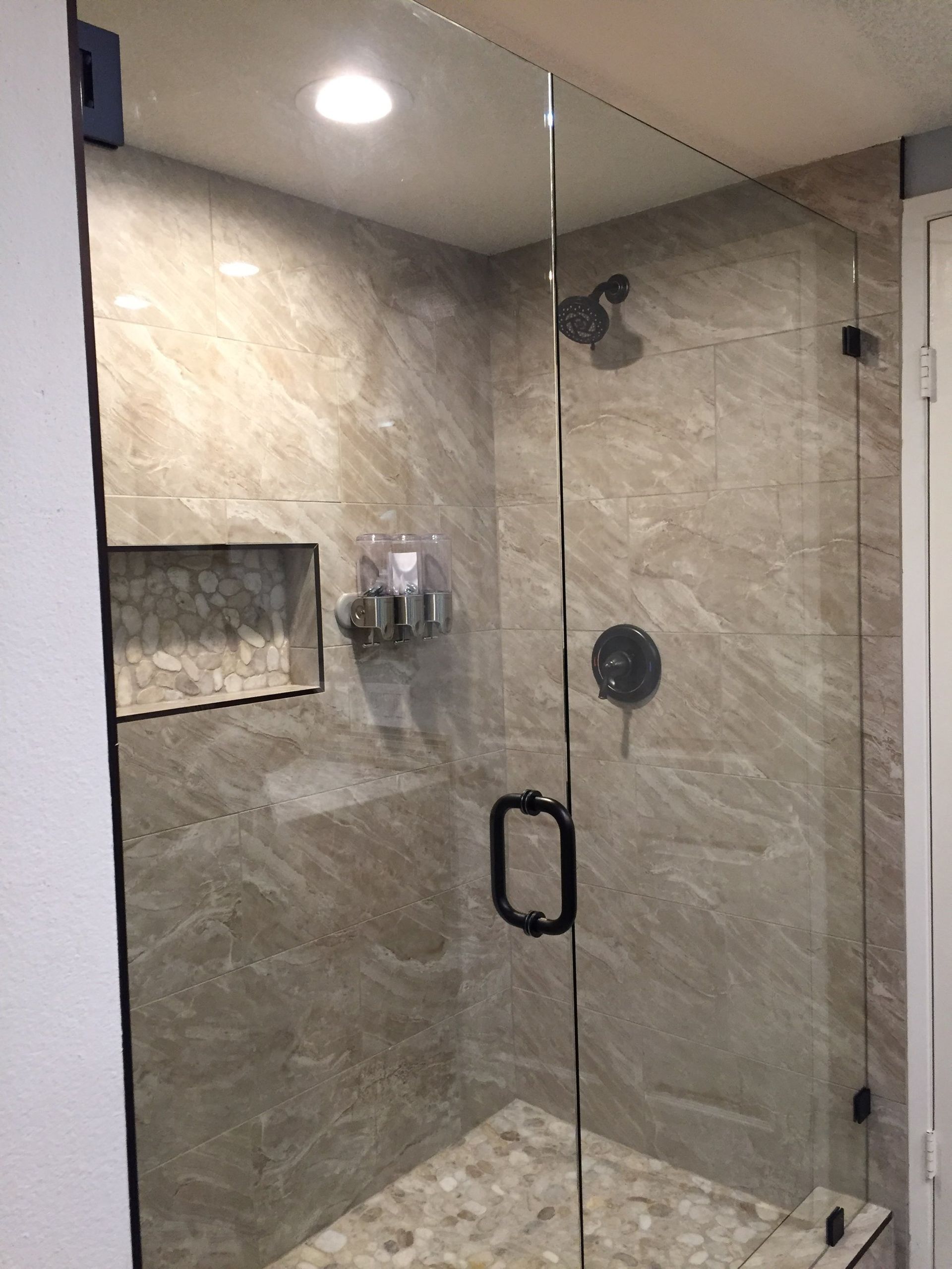 Modern glass shower with beige stone tile, recessed shelf, and chrome fixtures