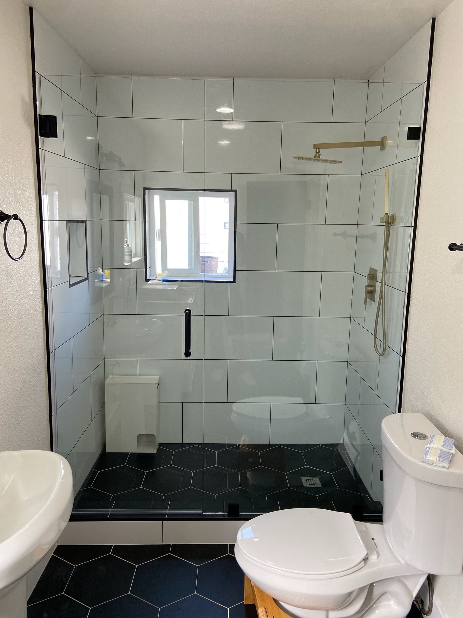 Small bathroom with white tiled walls, black speckled floor, toilet, sink, and a shower area with a window.