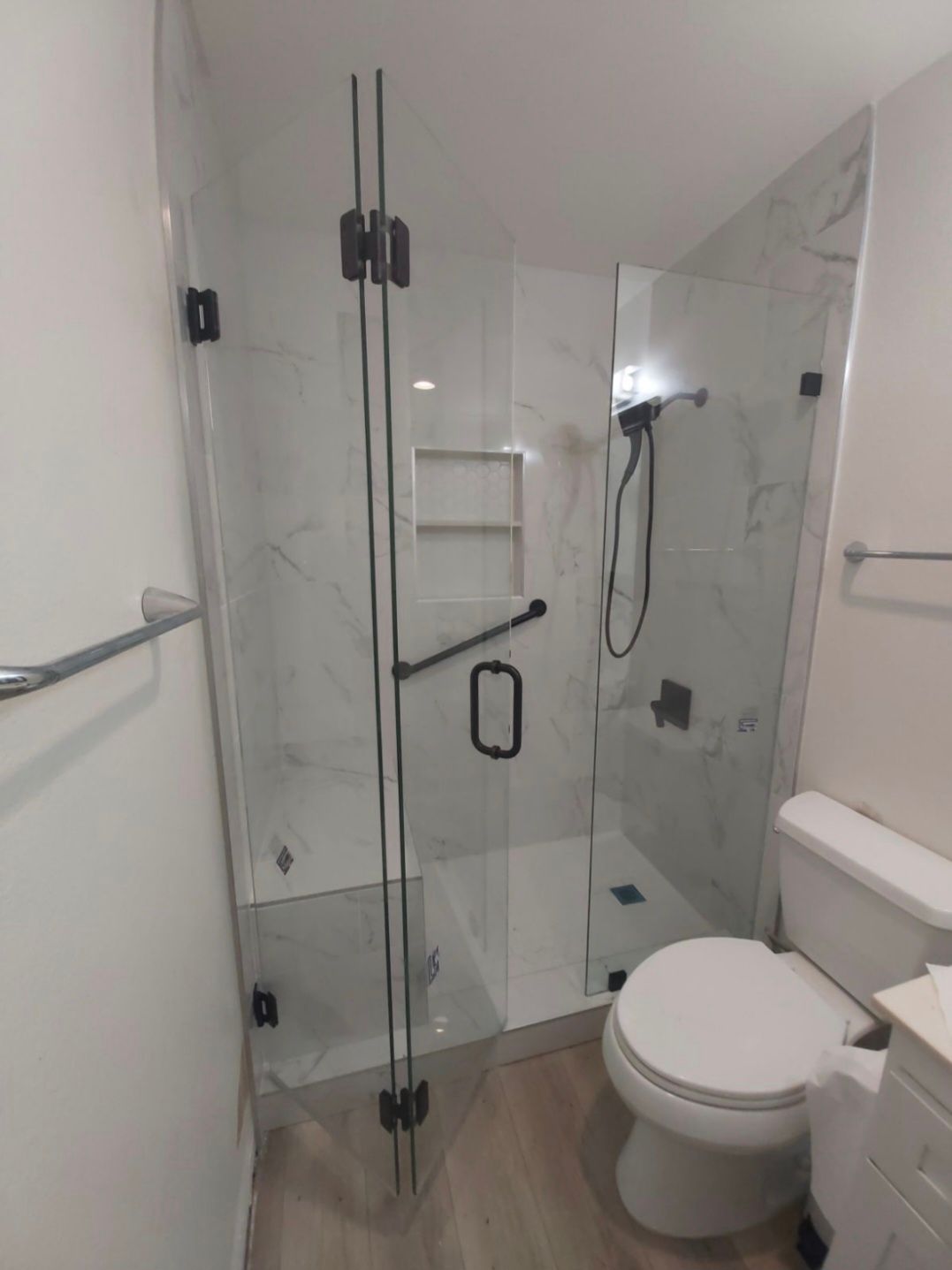 Modern bathroom with glass shower enclosure, toilet, white tile walls, and wood-look floor.