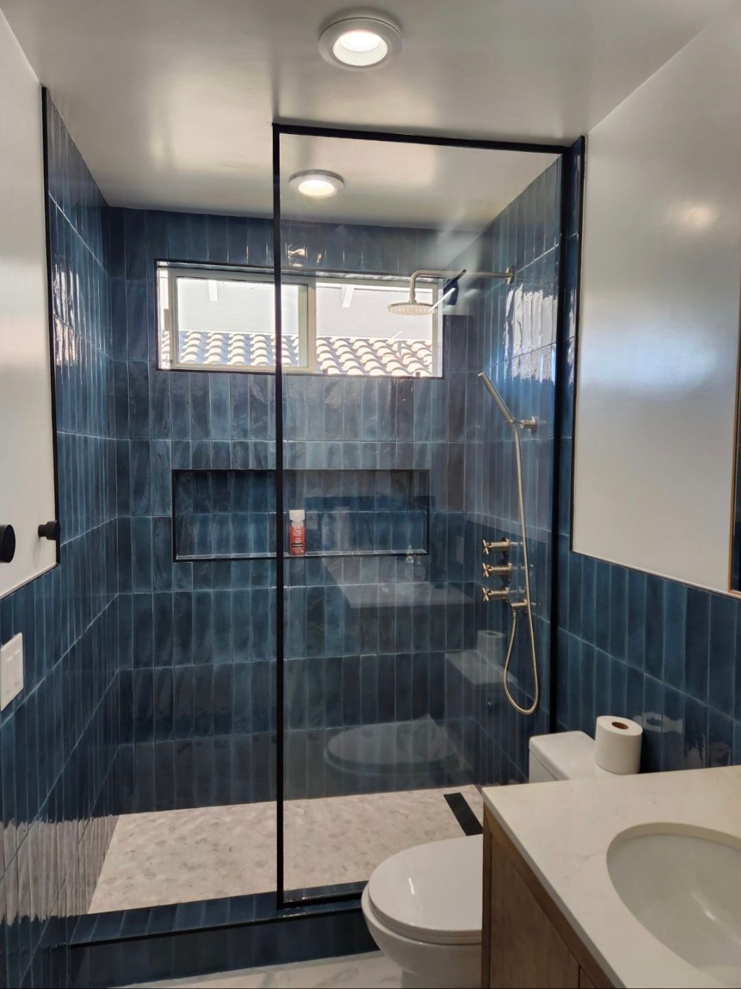 Blue-tiled bathroom with glass shower, toilet, and sink by a window