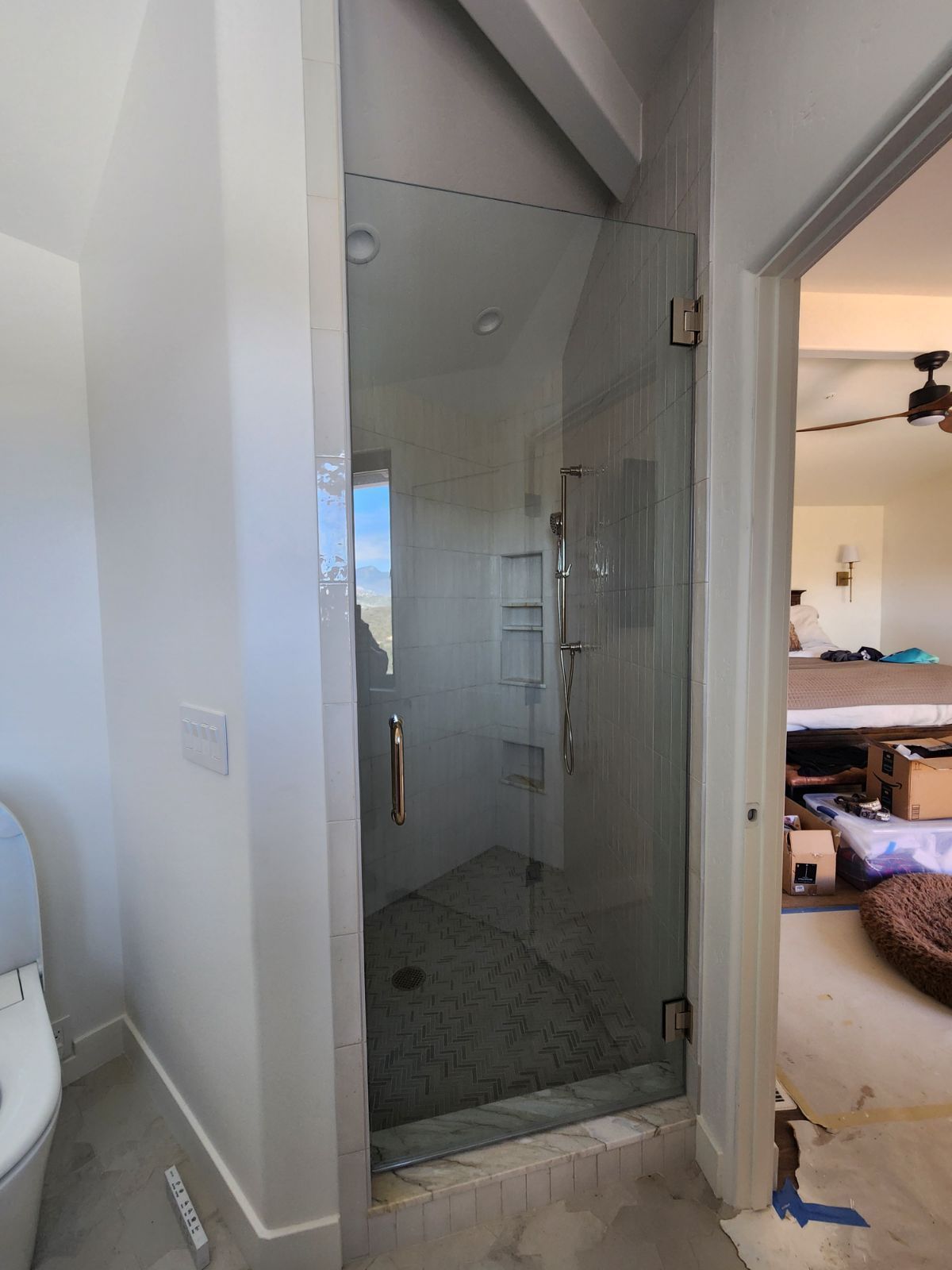 Glass shower stall in a bathroom, viewed through an open doorway, with tiled floor and ladder visible inside.