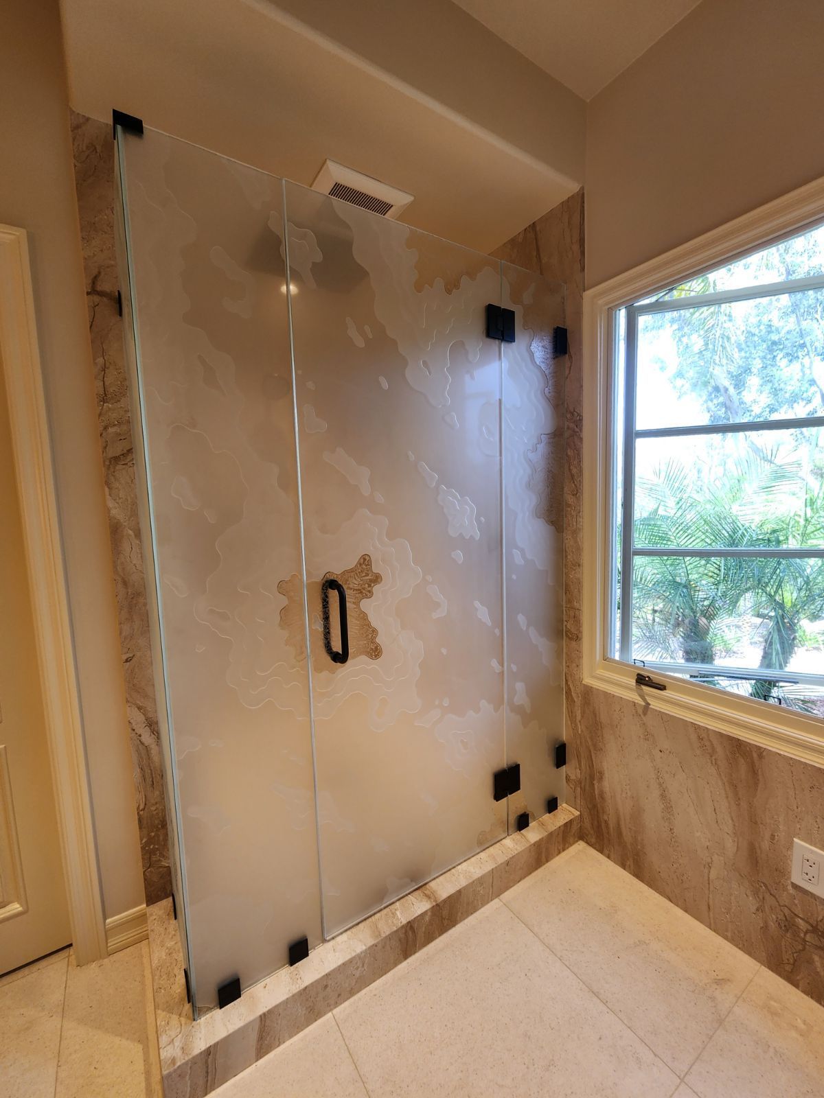 Glass shower enclosure in a beige bathroom beside a window, with tiled floor and walls.