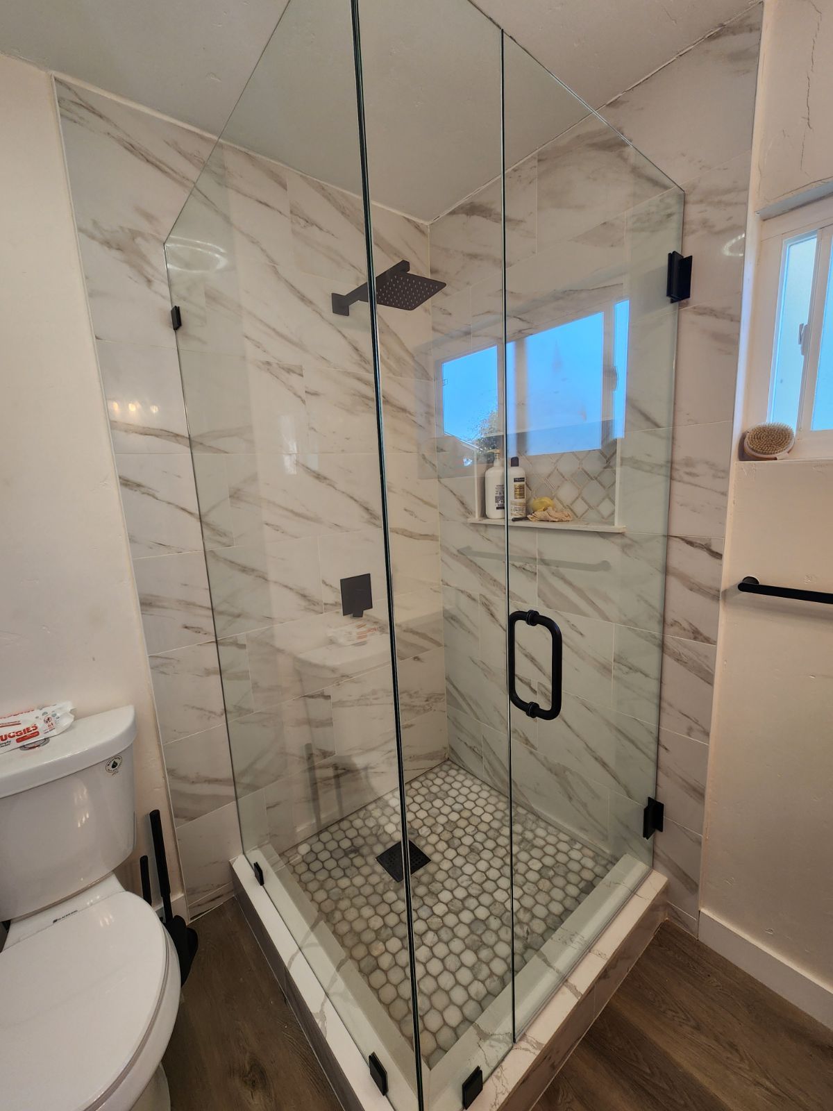 Modern bathroom shower with glass enclosure, white marble-look walls, hex tile floor, and a small window.