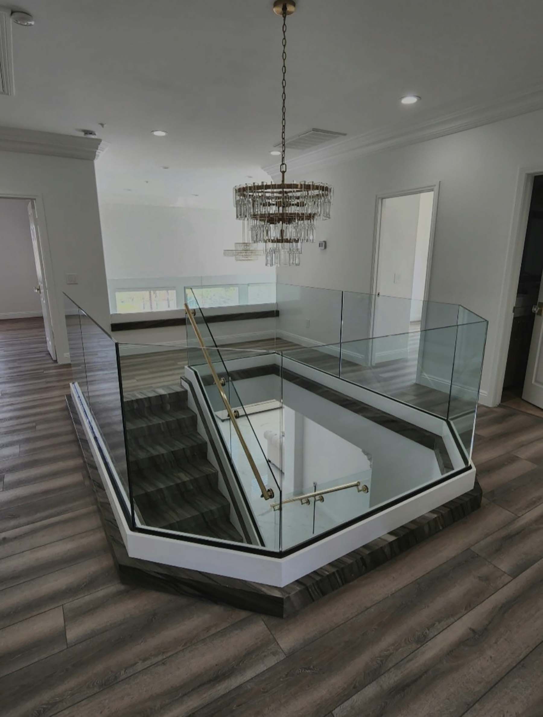 Modern home foyer with glass-railed staircase and chandelier, viewed from the landing.