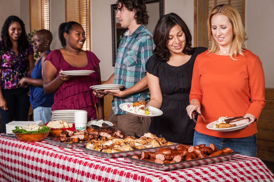 People at a buffet, serving themselves. Red checkered tablecloth with meat, salad. Indoors, cheerful expressions.