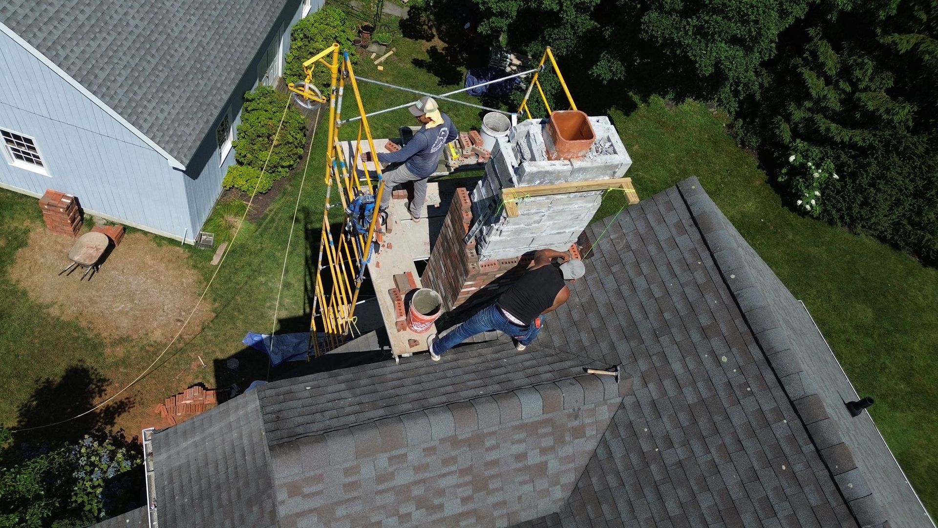 An aerial view of two workers on a scaffold rebuilding a stone chimney on a gray-shingled roof.