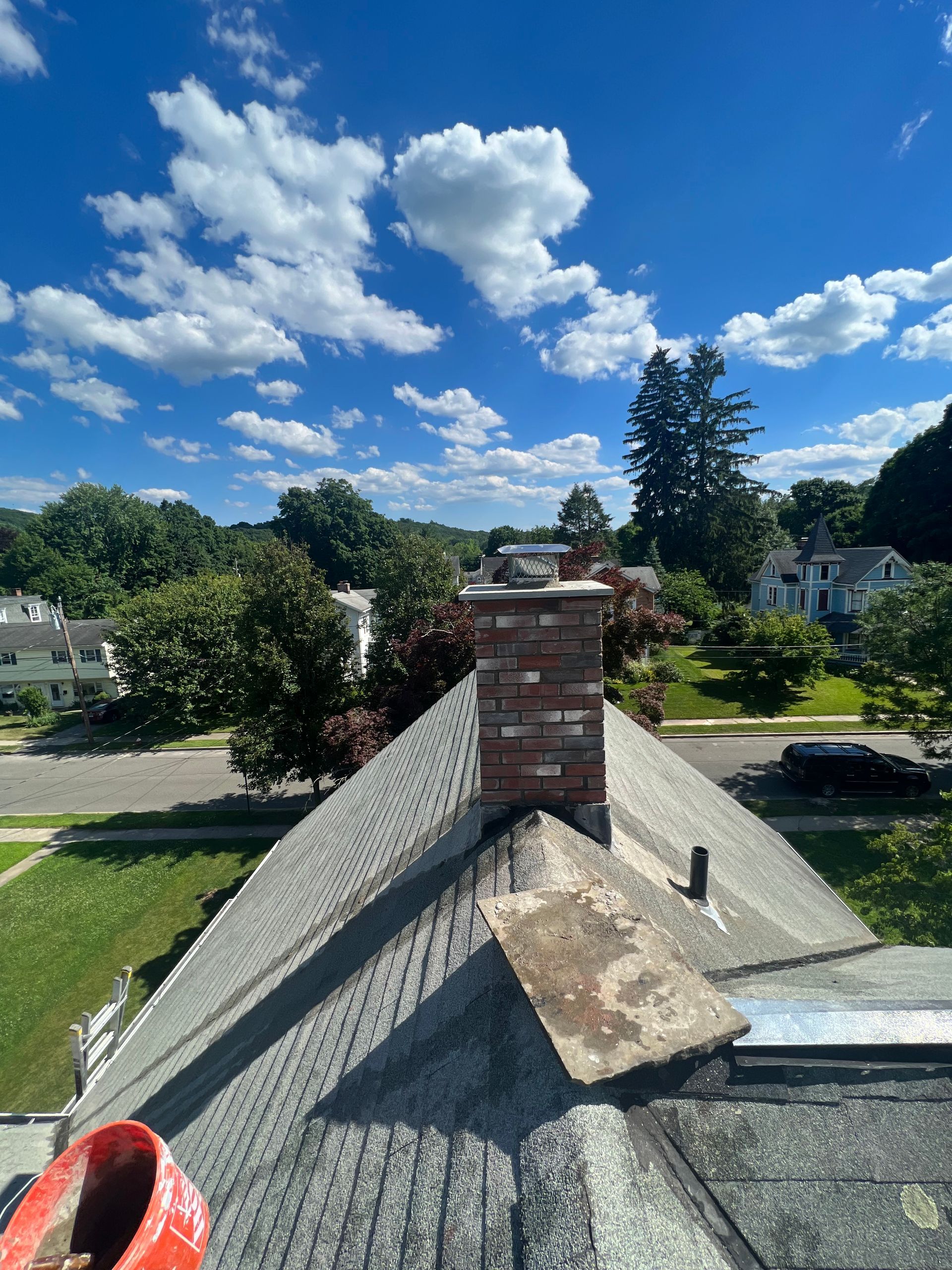 A brick chimney stands atop a grey shingled roof under a bright blue, cloudy sky, with a neighborhood visible below.