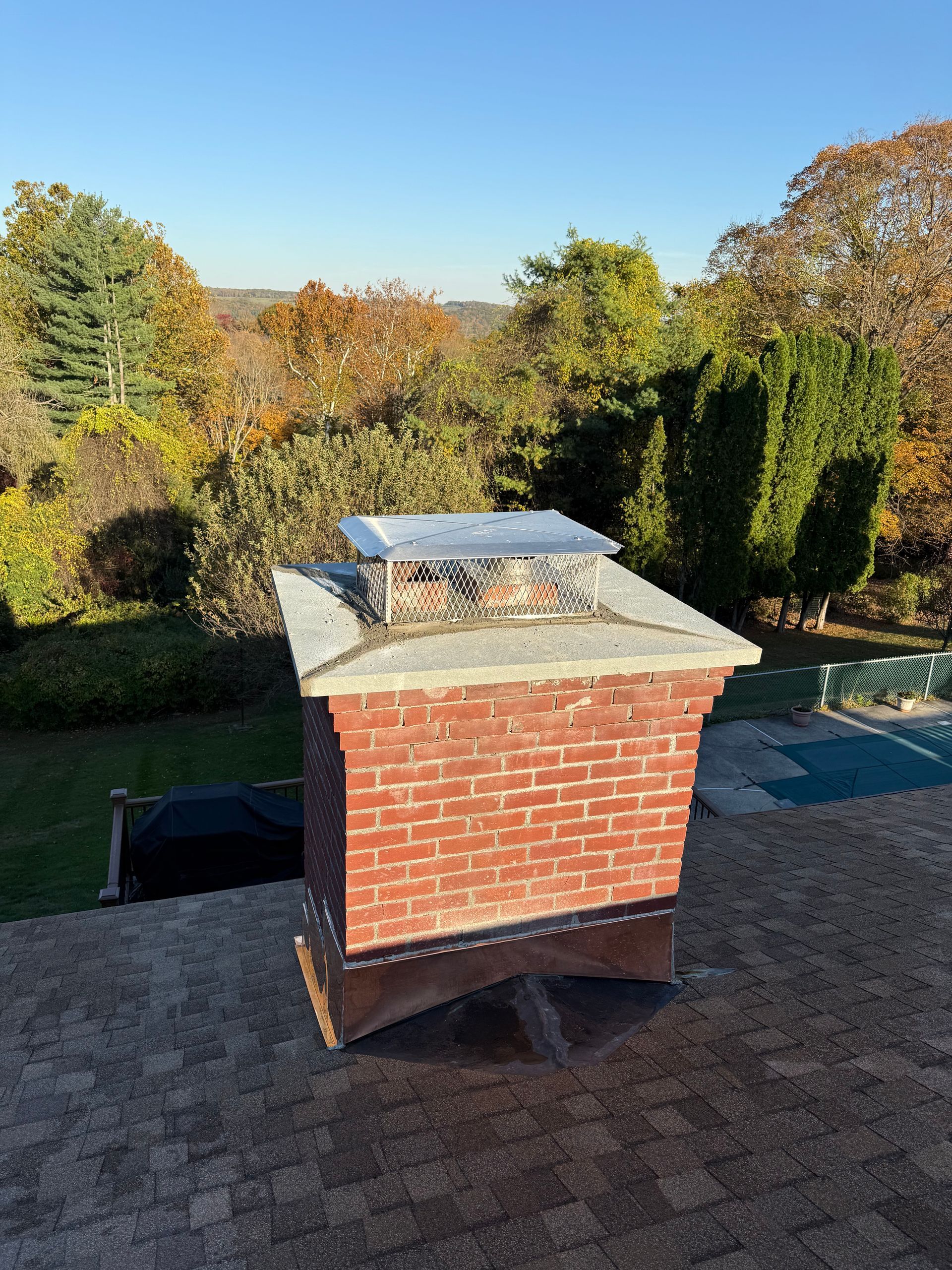 A brick chimney with a metal cap and copper flashing stands on a shingled roof, set against a backdrop of autumn trees.