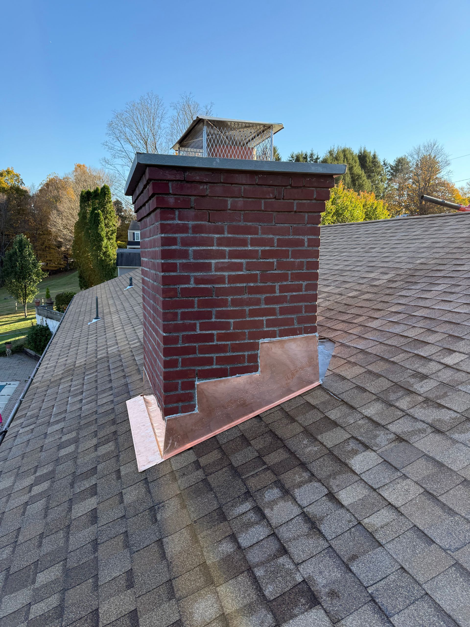 A red brick chimney on a shingled roof features newly installed copper flashing around its base against a blue sky.