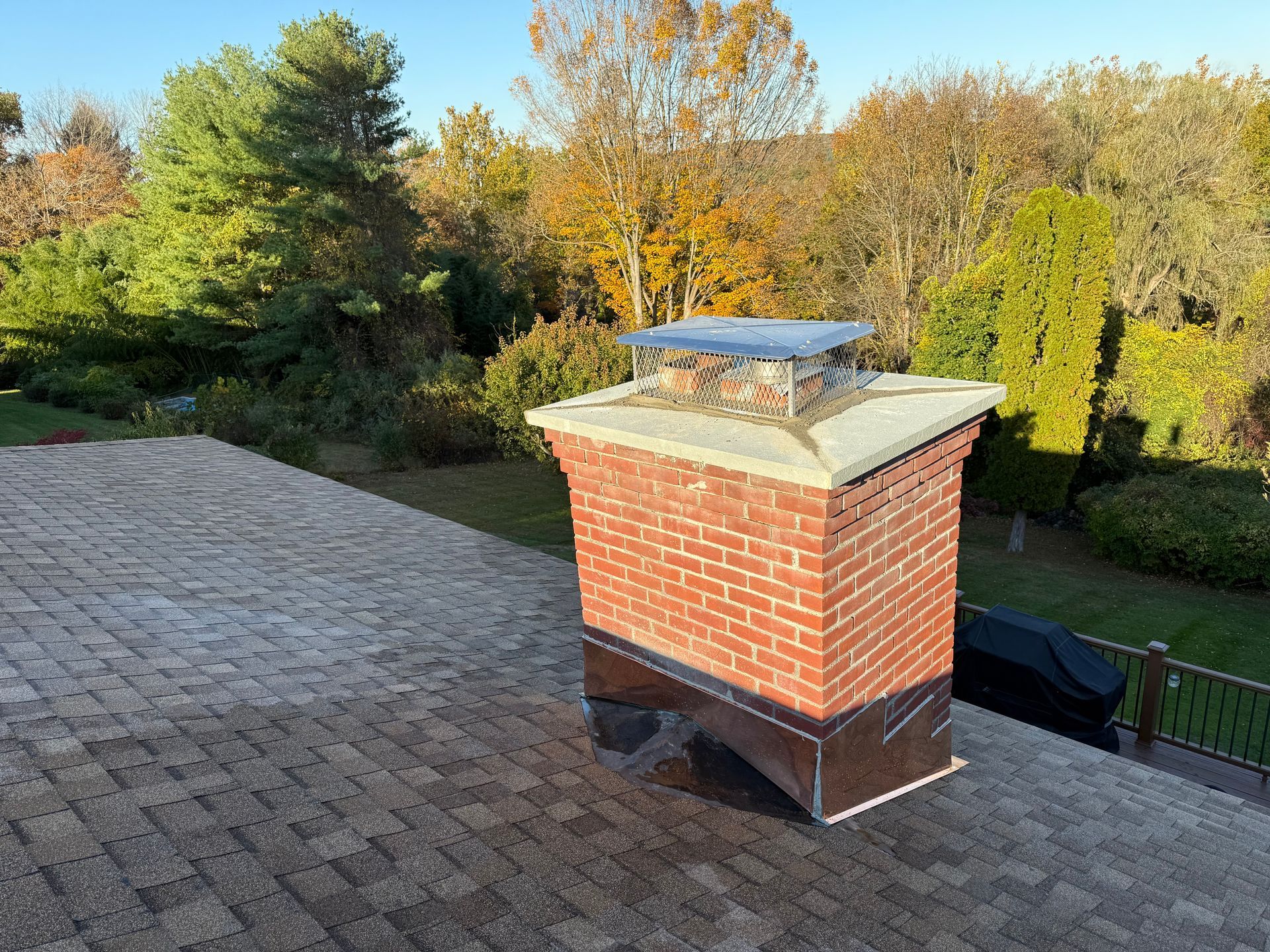 A brick chimney with a metal cap stands on a shingled residential roof surrounded by trees in autumn.