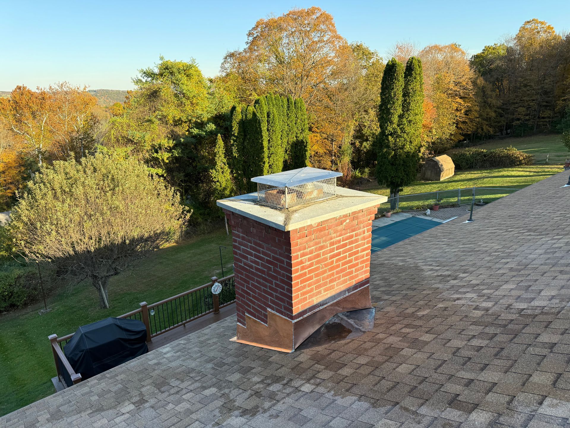A brick chimney with a metal cap stands on a paved stone roof, overlooking a wooded yard with a pool.