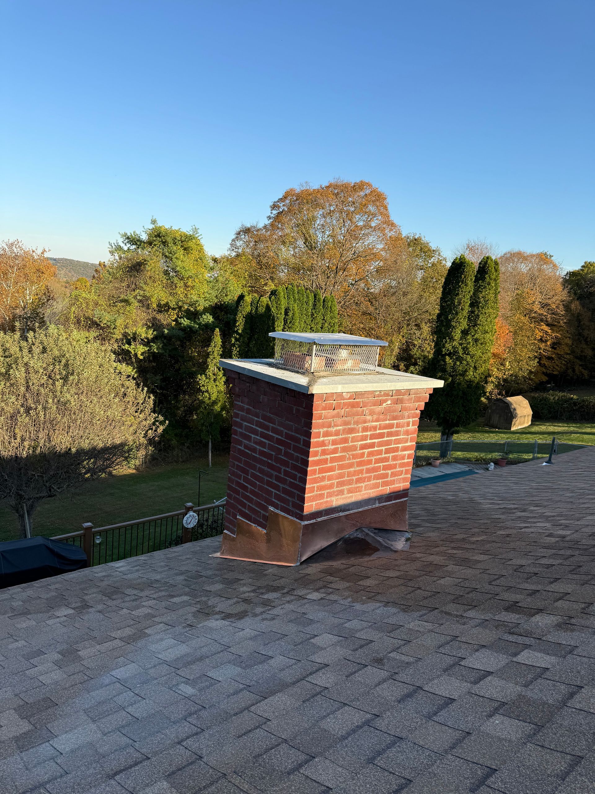 A red brick chimney with a metal cap sits on a shingled roof against a backdrop of trees under a clear blue sky.