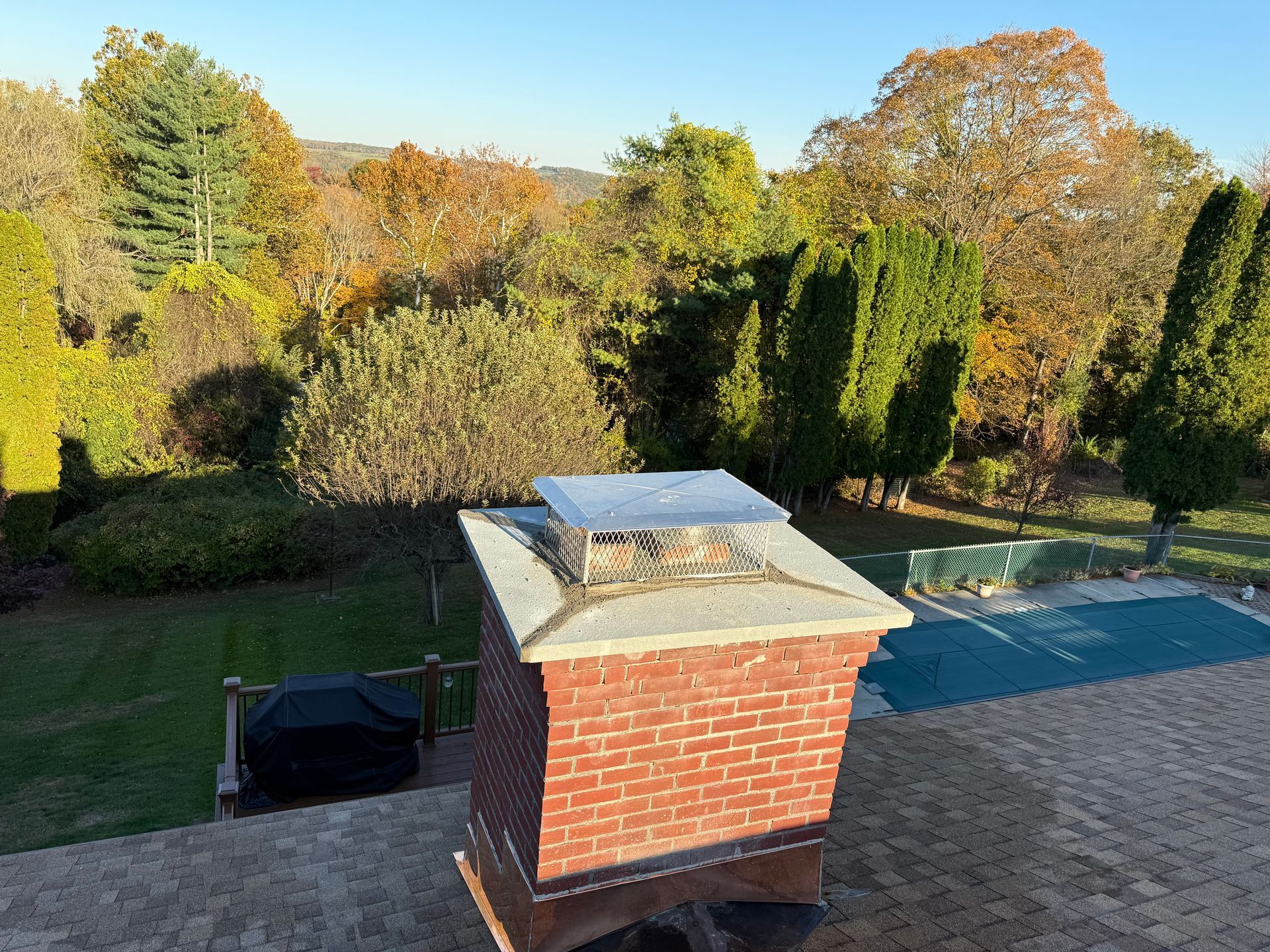 A metal chimney cap sits atop a red brick chimney on a shingled roof overlooking a green yard and swimming pool.