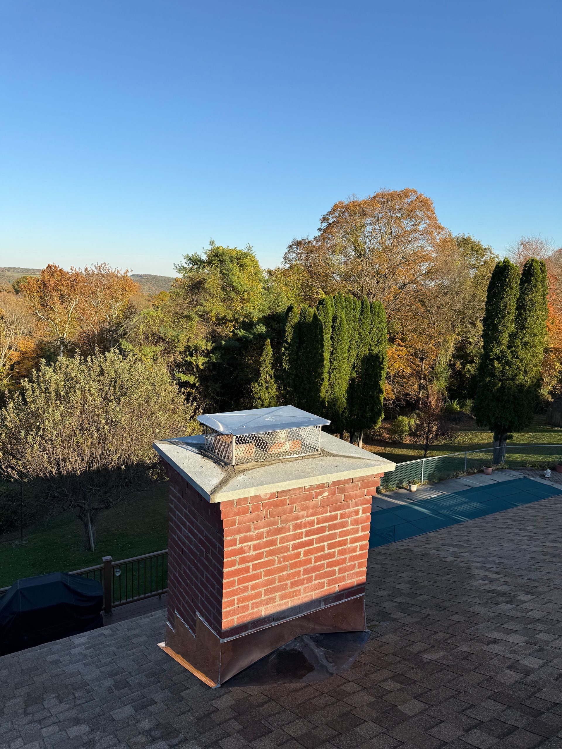 A brick chimney with a metal cap stands on a shingled roof, set against a background of autumn trees and a blue sky.