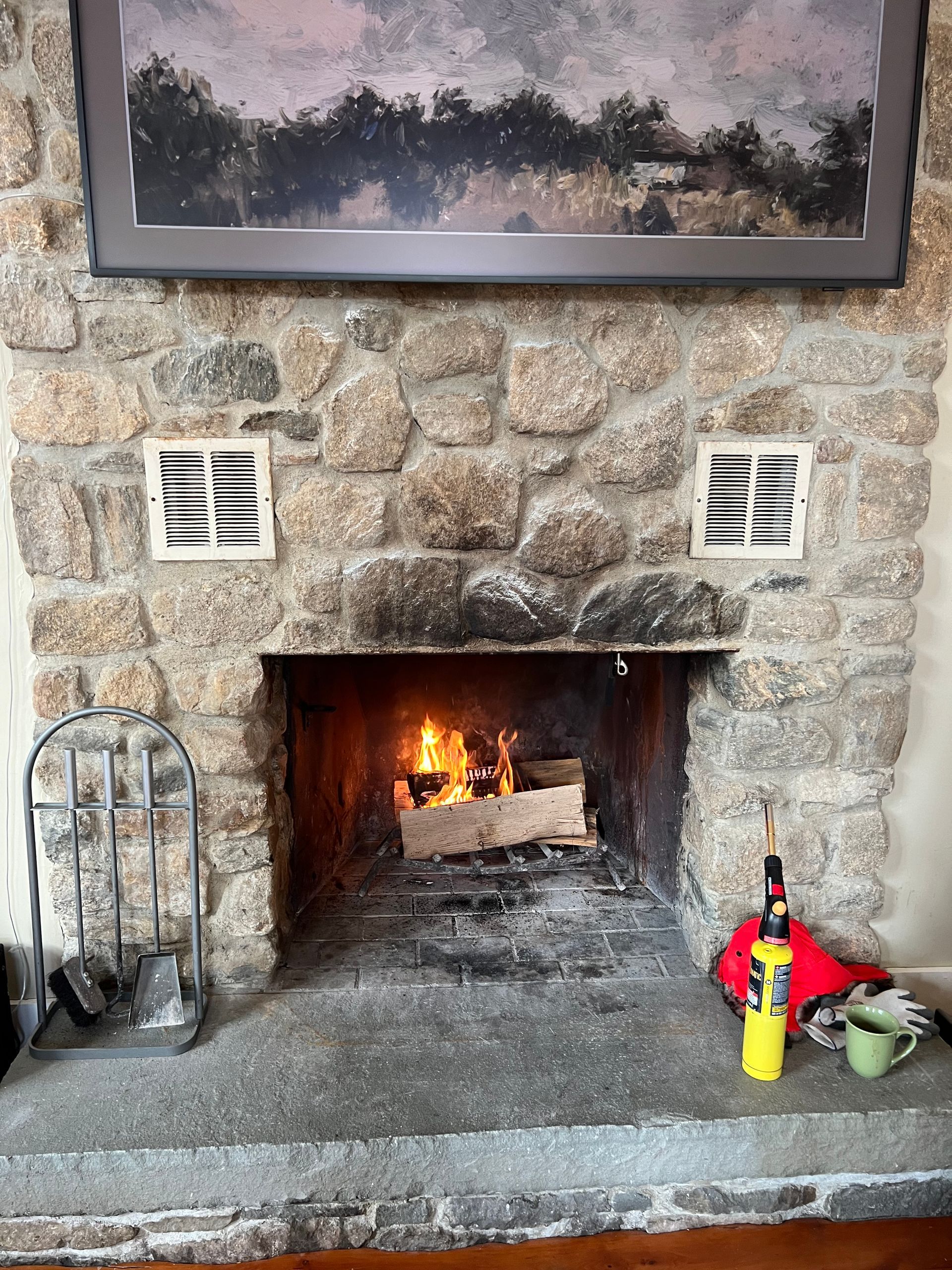 A stone fireplace with a burning fire, a metal tool set, and a yellow canister on the hearth beneath a framed painting.