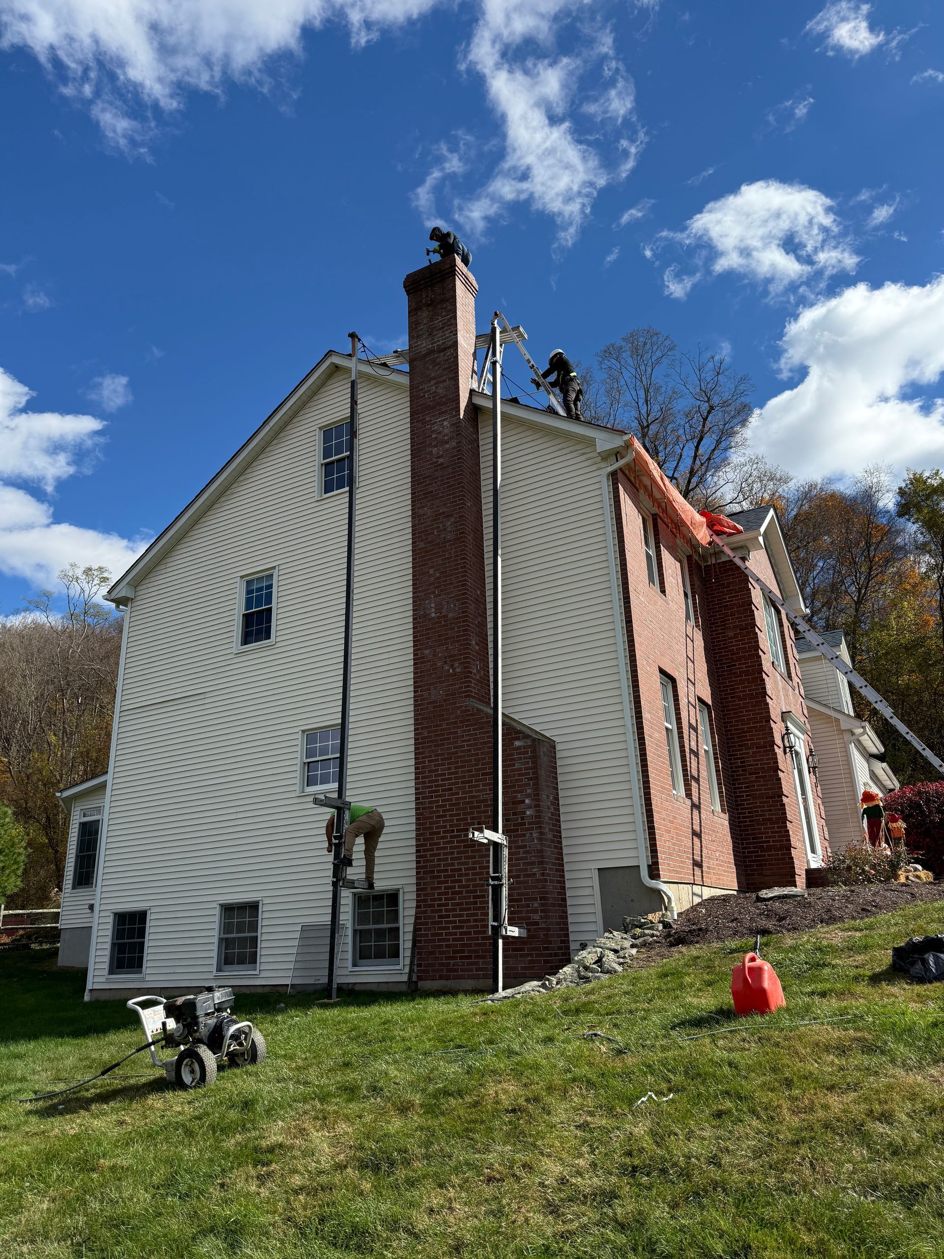 Workers on the roof of a two-story home with light siding and a brick chimney during a sunny day.