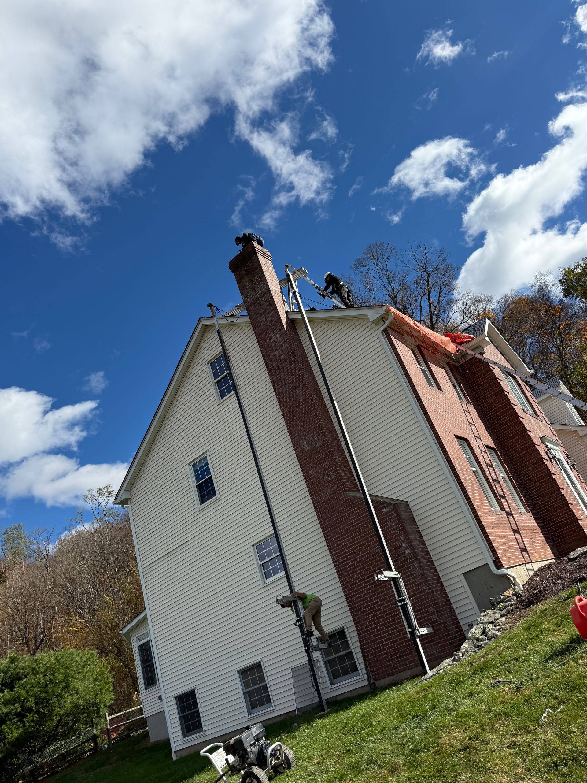 Roofers work on a house with a brick chimney and light-colored siding under a blue sky with scattered clouds.