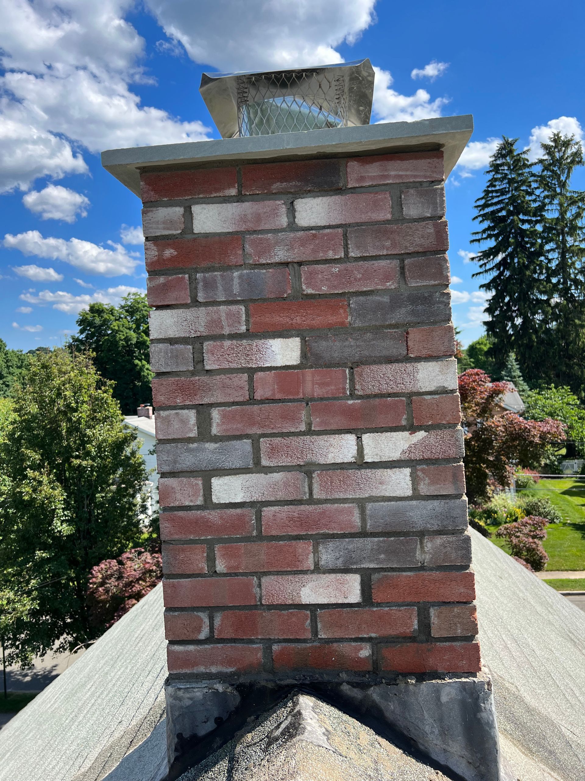 A brick chimney on a roof under a bright blue sky with trees and neighborhood houses in the background.