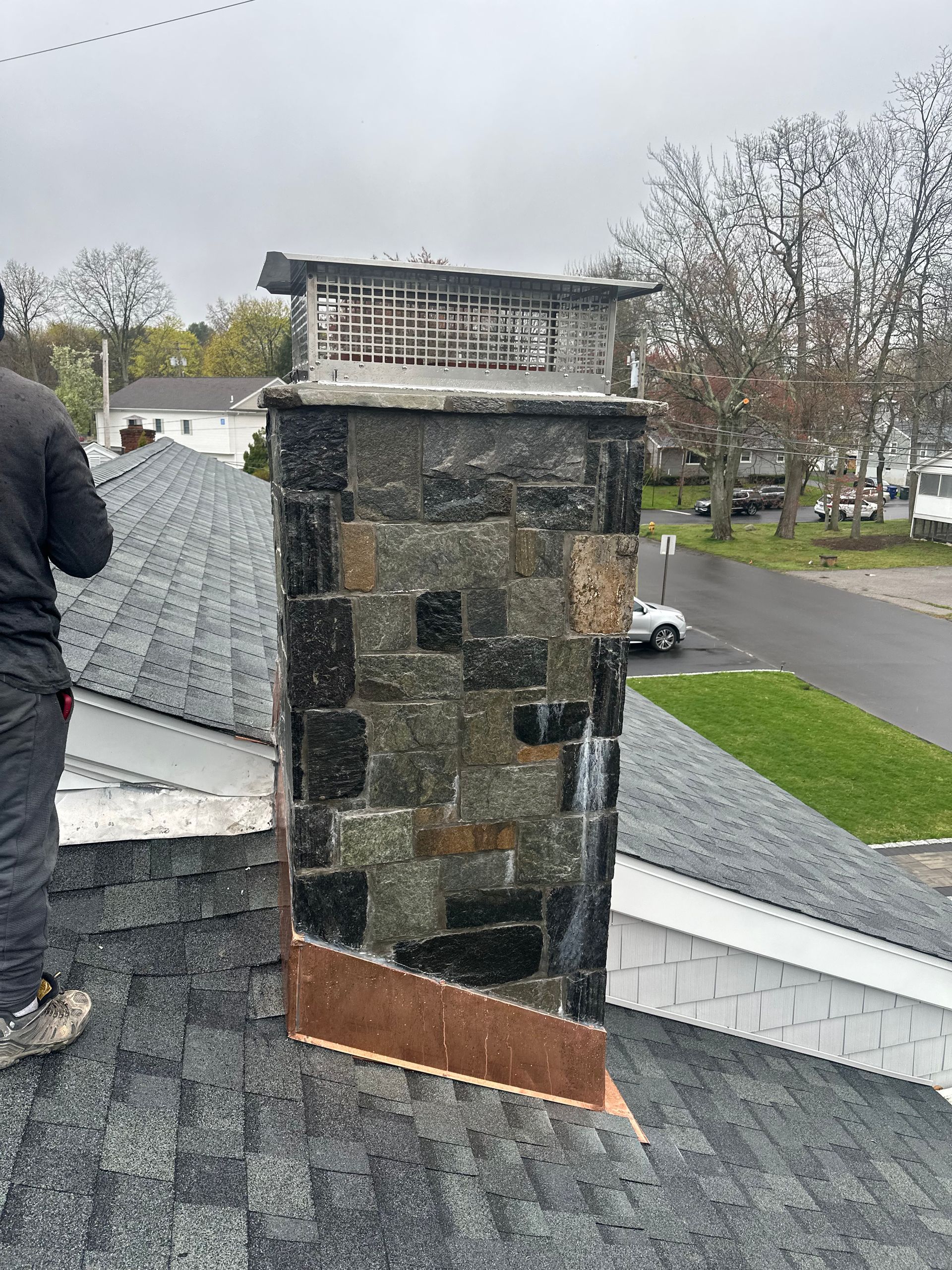 A stone chimney with a metal cap on a residential roof, featuring copper flashing at the base and a worker to the left.