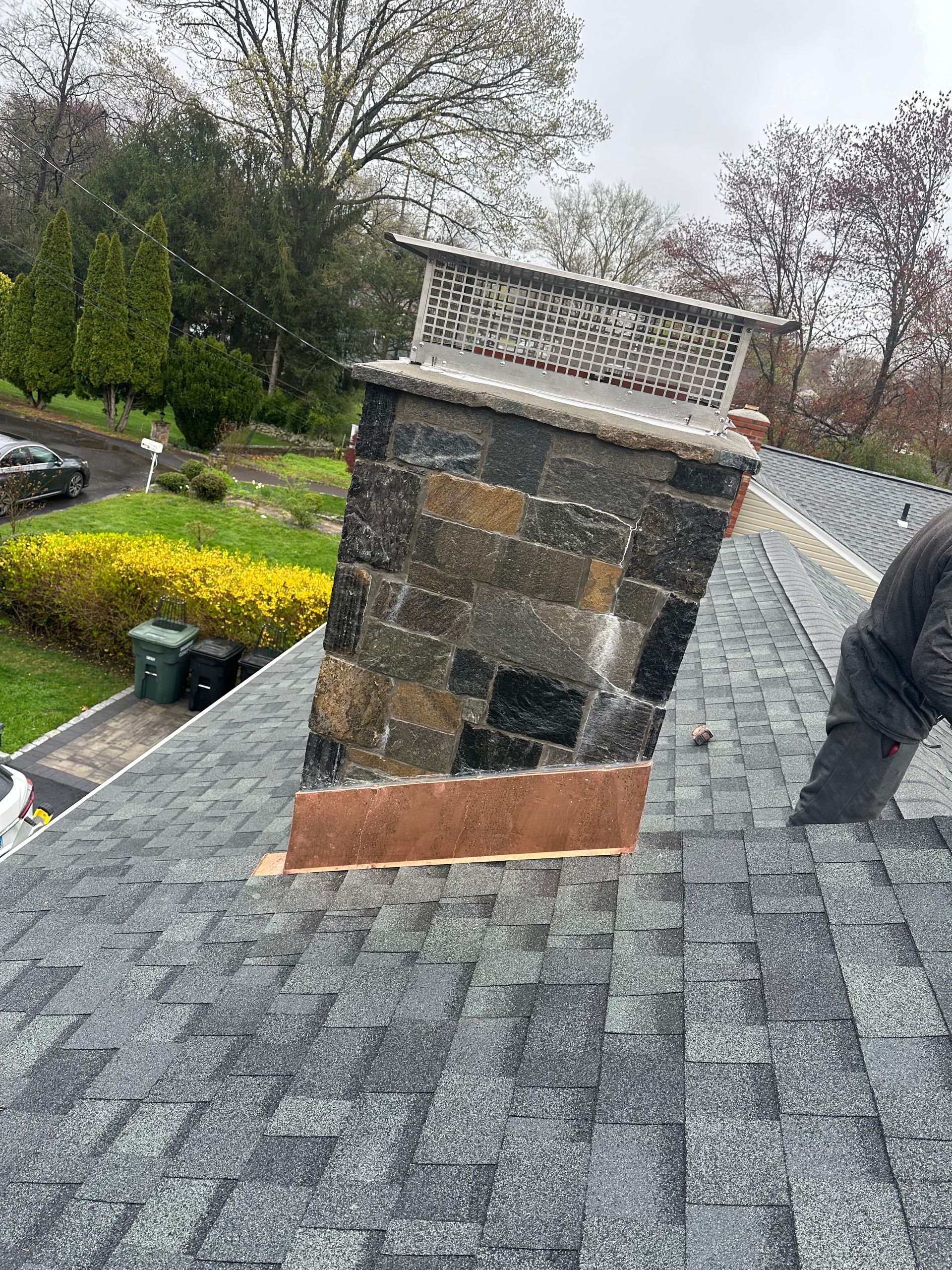 A stone chimney with a decorative metal cap stands on a sloped shingled roof with a person visible on the right.