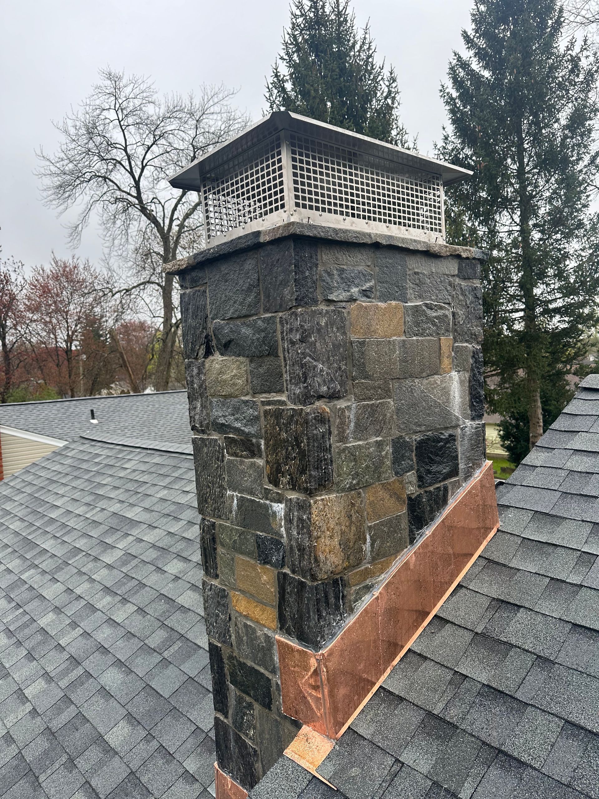 A stone chimney on a residential roof, featuring a metal cap and copper flashing at the base against shingles.