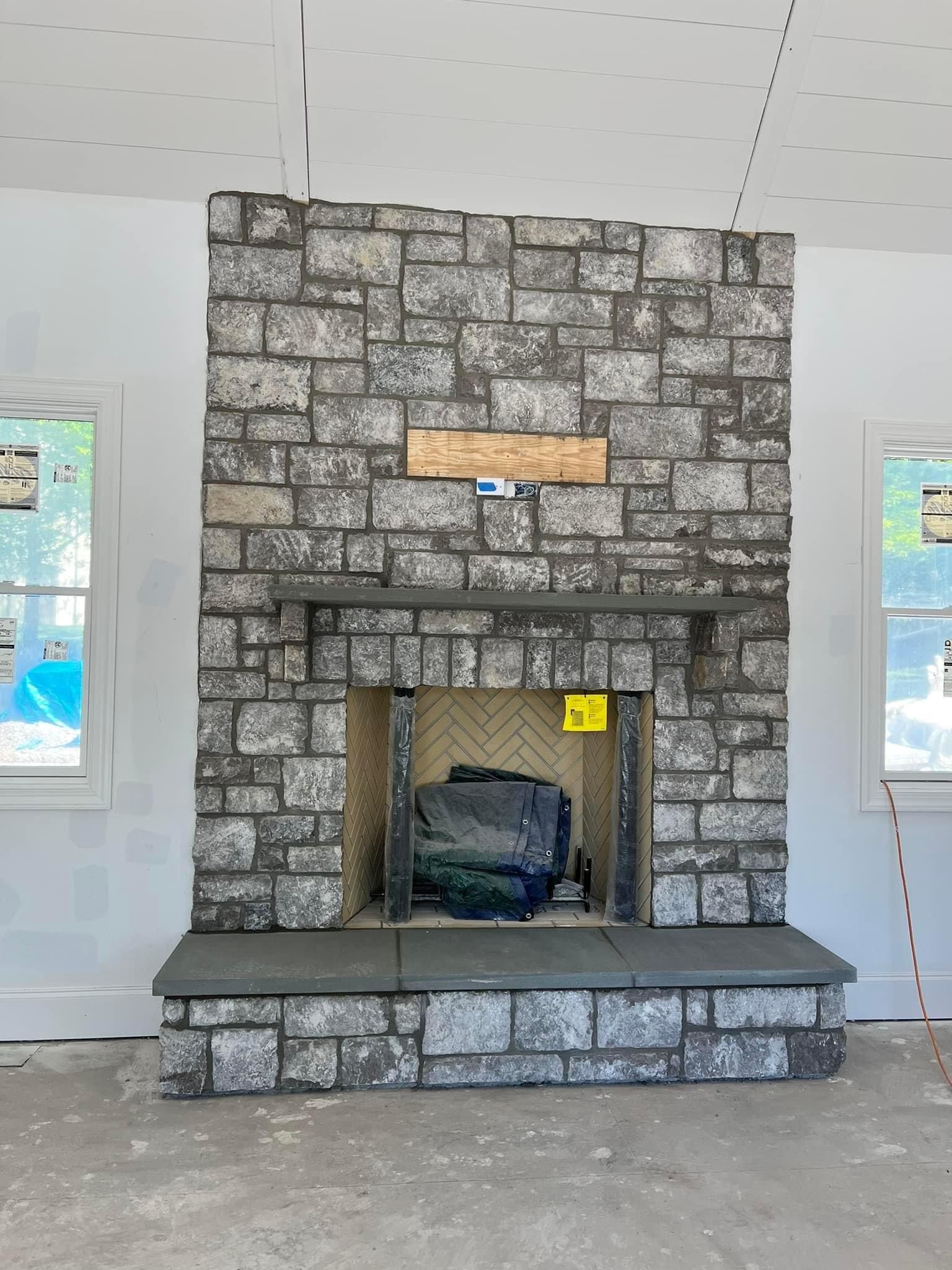 A stone fireplace under construction, featuring gray masonry, a dark hearth, and a wooden mantel insert in a room.