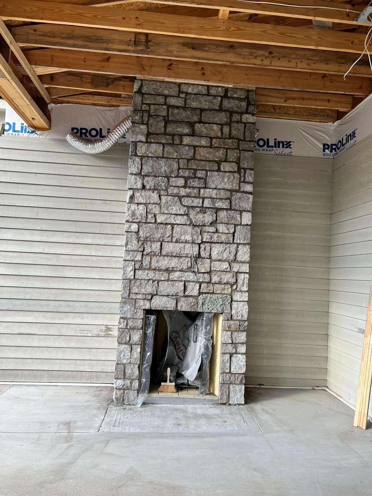 A stone fireplace under construction in a room with exposed ceiling joists and unfinished wall sheathing.